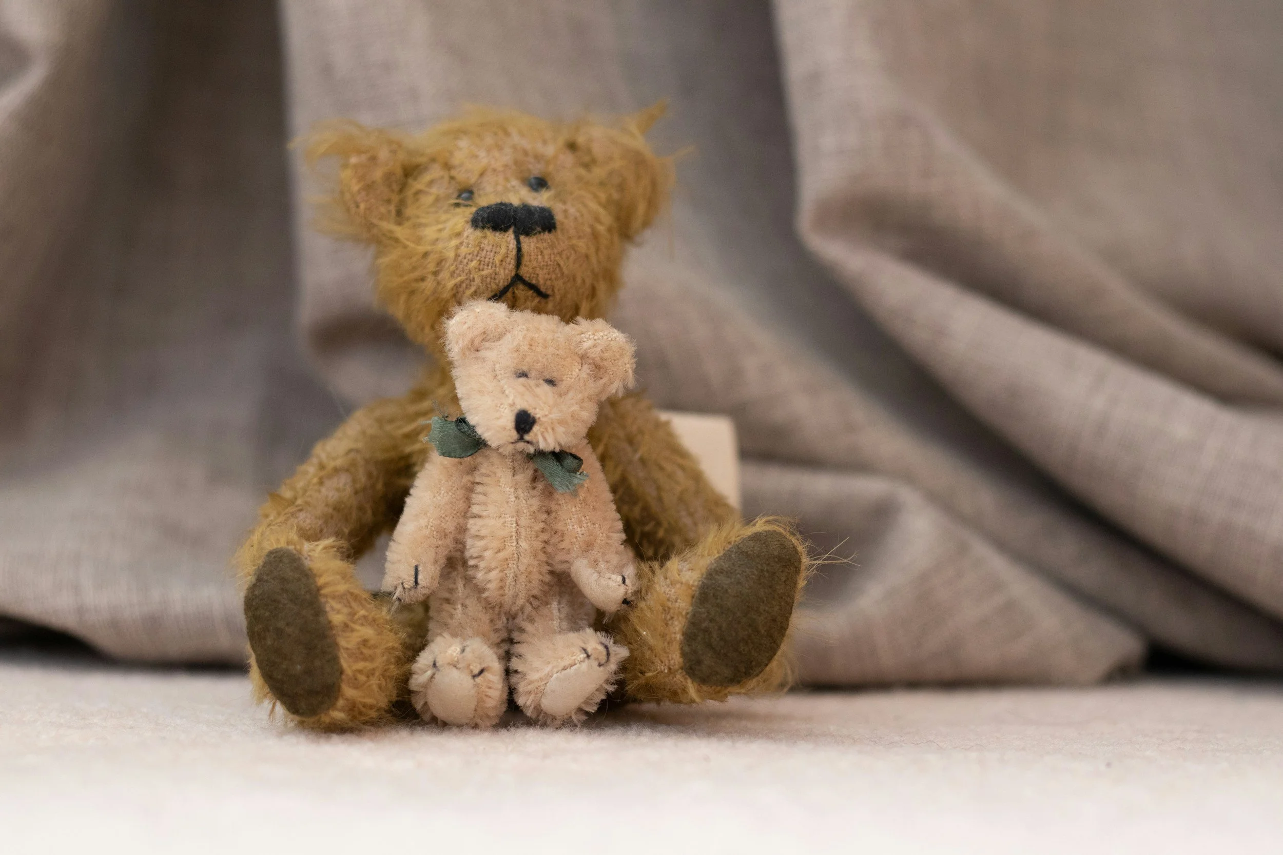 Two teddy bears, one larger and one smaller, sit in front of a gray fabric background. The larger teddy bear is brown with a black nose and eyes, and the smaller teddy bear is a lighter color with a green bow around its neck.