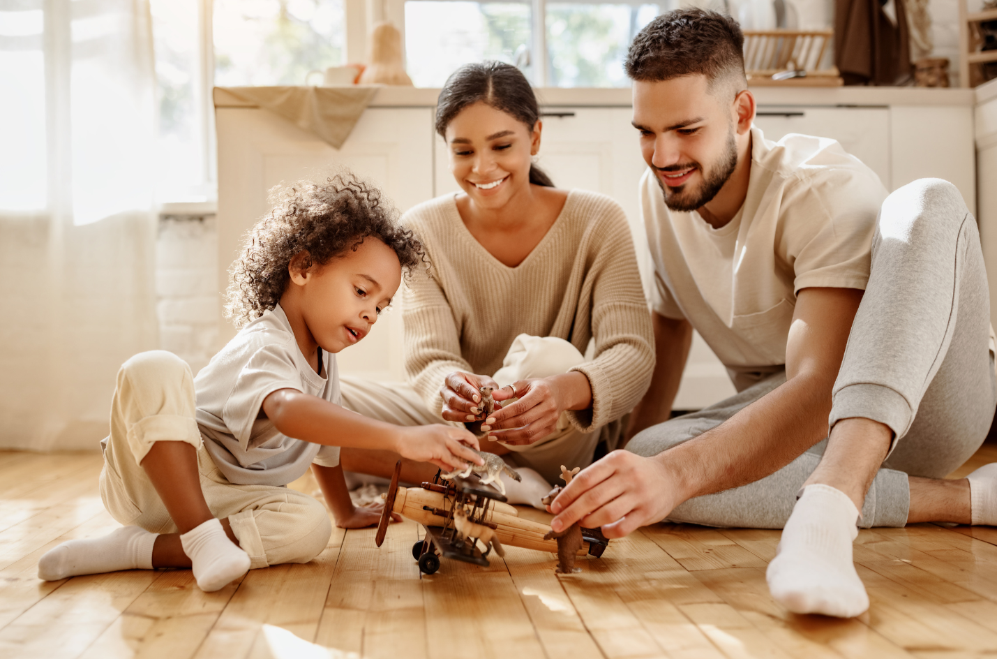 A family of three playing with toy airplanes on a hardwood floor in a sunlit kitchen, smiling and engaged in their activity.