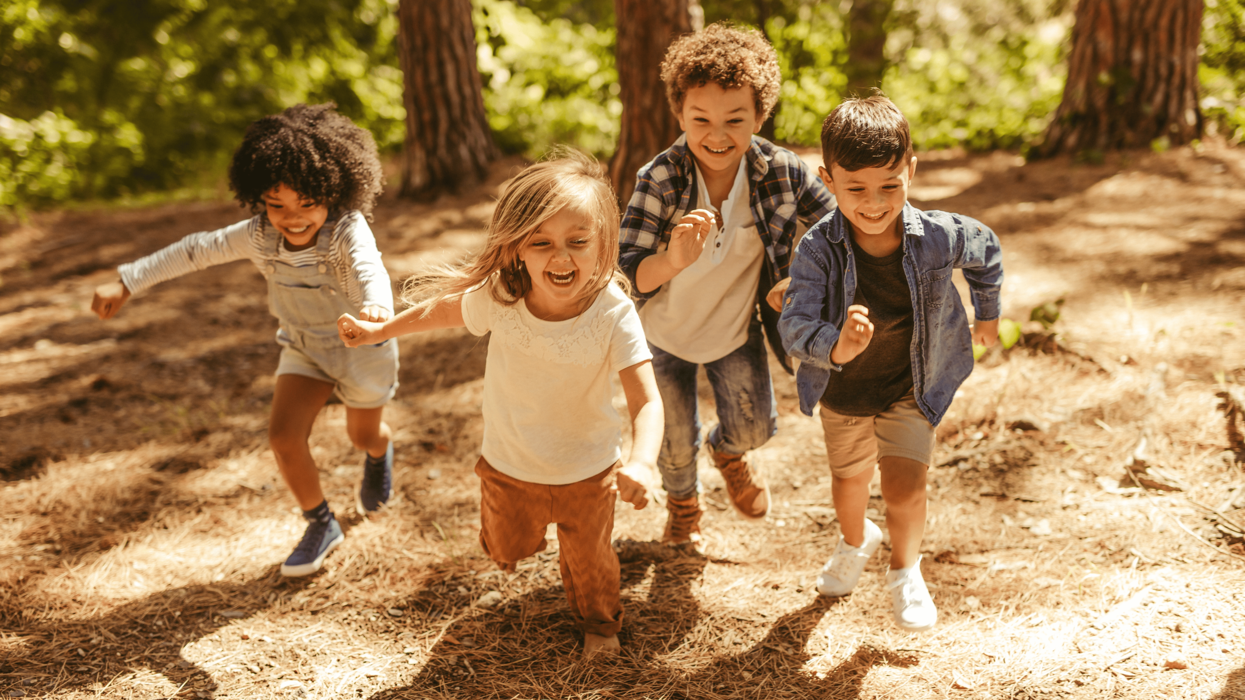 Group of five children running and playing outdoors in a wooded area with sunlight filtering through trees.