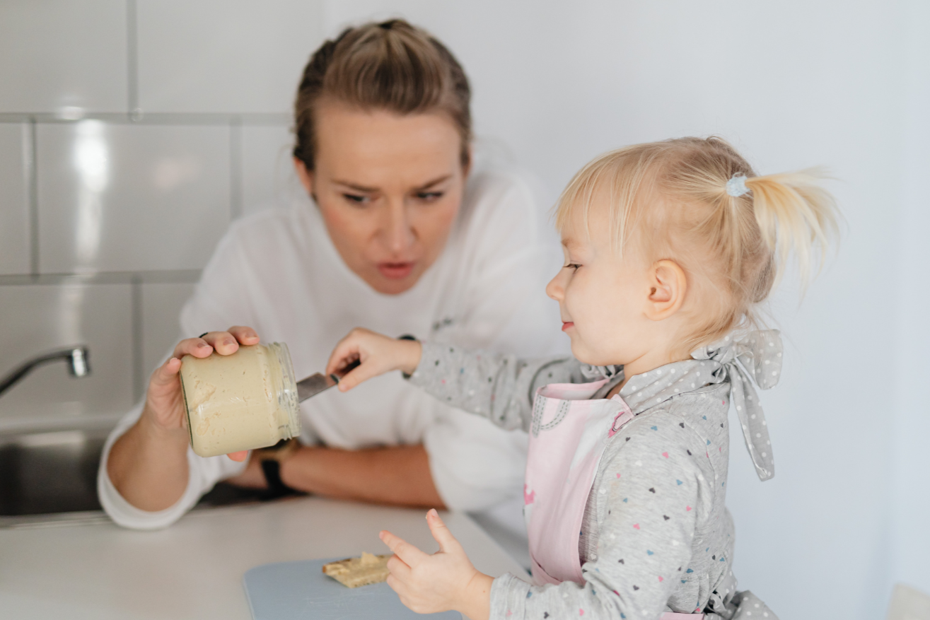 A woman and a young girl in a kitchen with gray tiled wall, preparing a snack. The girl is spreading peanut butter on bread, smiling, while the woman looks on with a surprised expression.