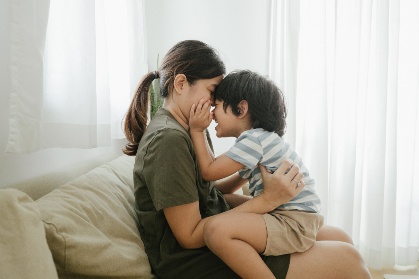 A woman and a young boy touching their foreheads together in a loving moment in a bright living room.