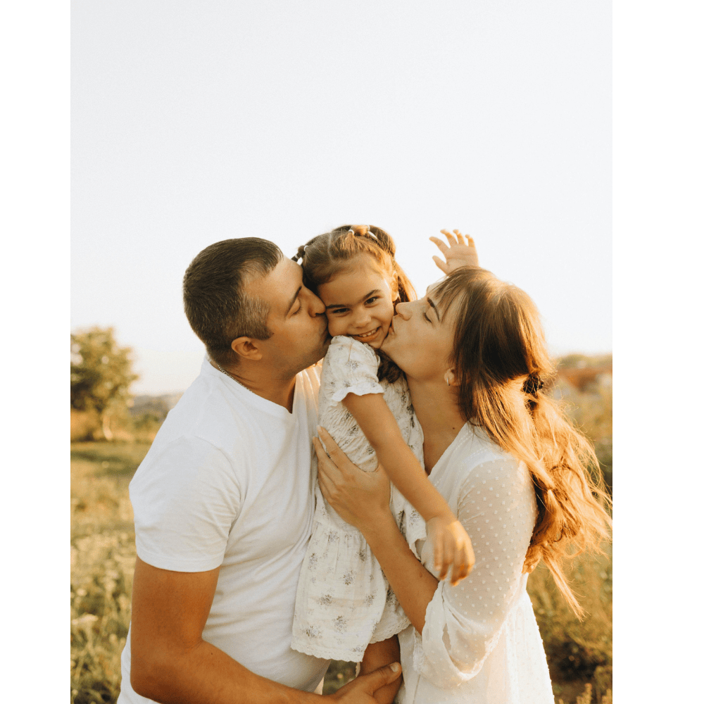 Family of three outdoors, father and mother kissing their young daughter on the cheeks, happy and smiling, in a field during golden hour.