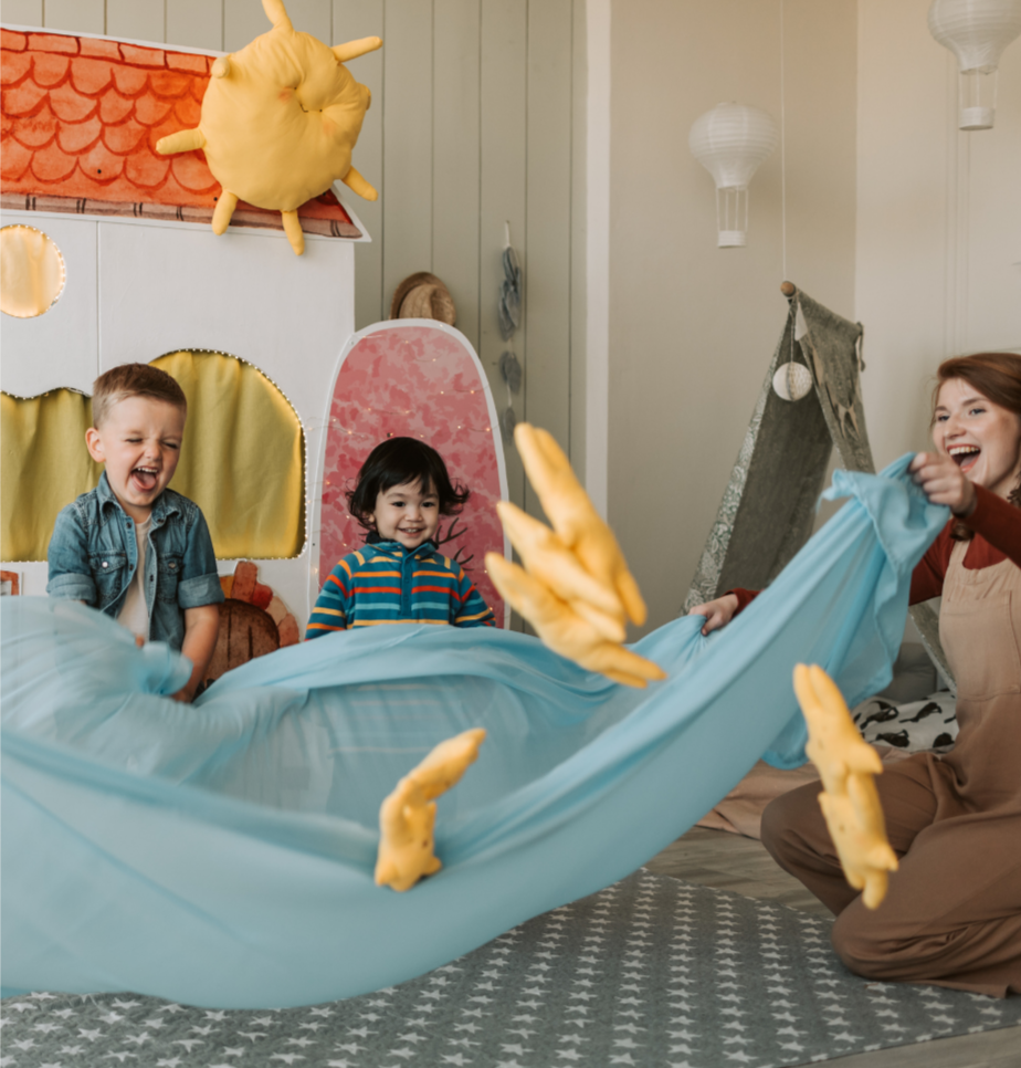 Children playing with a blue fabric and yellow plush toy hands in a decorated playroom with paper lanterns.