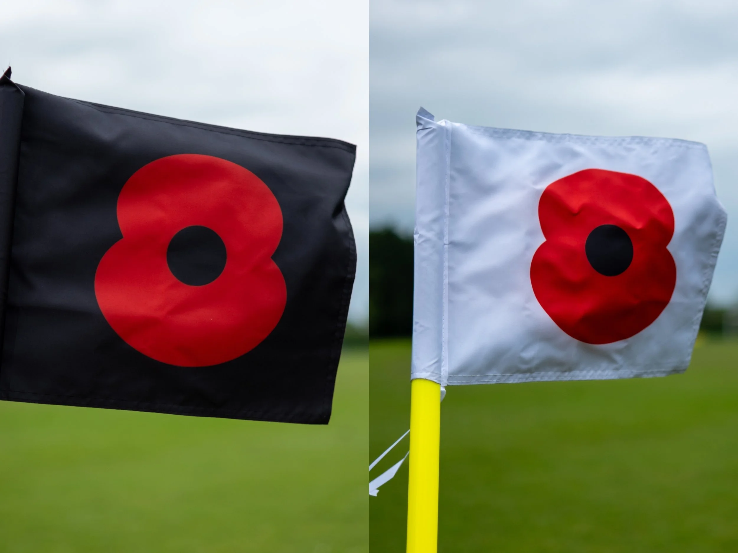 Close-up of two flags, one black and one white, each with a large red poppy flower symbol, flying outdoors on a cloudy day with a grassy field in the background.