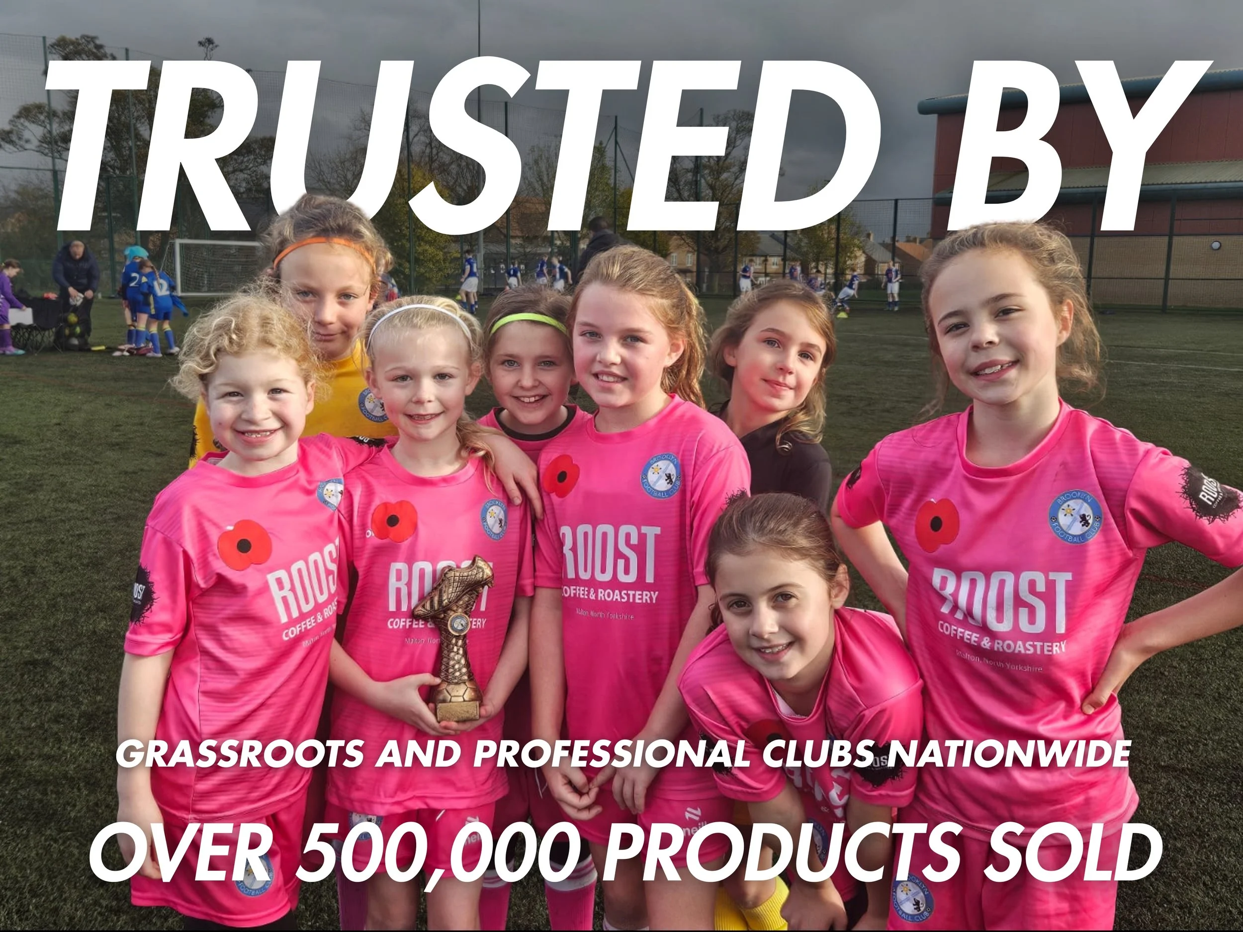 Group of young girls in pink soccer uniforms celebrating after a game on a soccer field, one holding a trophy.