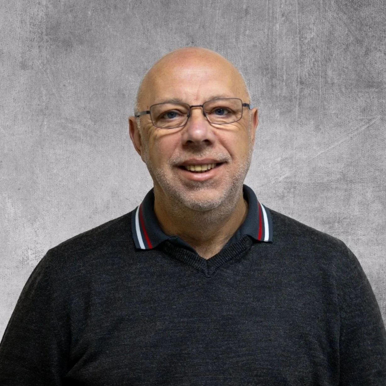 A smiling middle-aged man with glasses and a short beard, wearing a dark sweater with a collared shirt underneath, standing against a plain gray background.