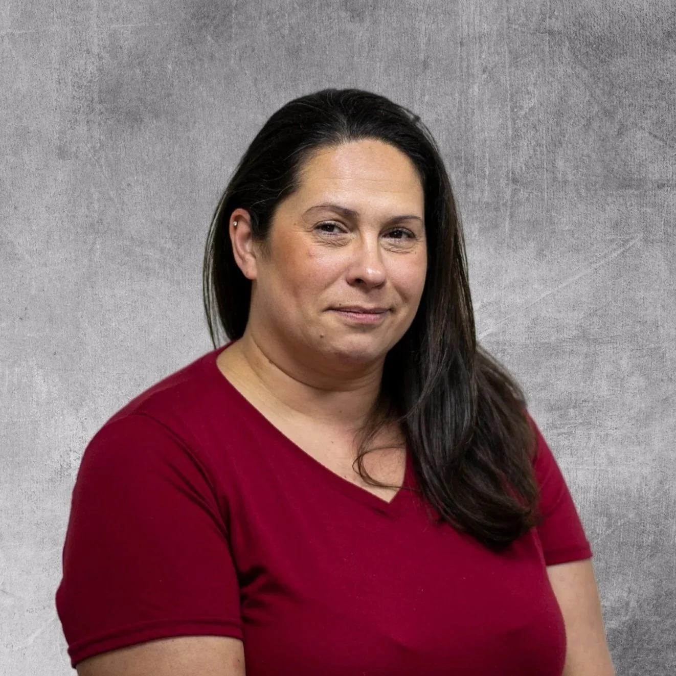 A woman with dark brown hair wearing a red shirt standing against a gray textured background.