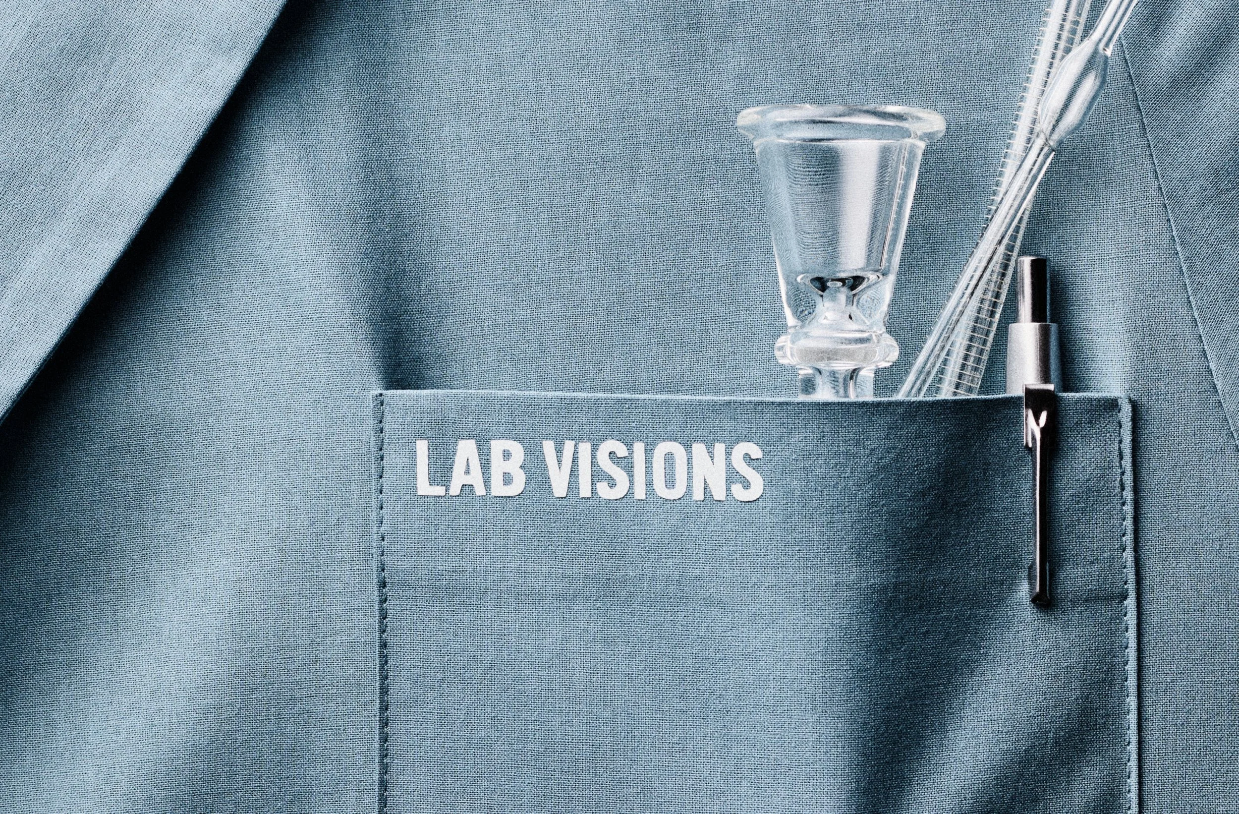 Close-up of a scientist's pocket on a blue lab coat, with glass pipette and other lab tools inside the pocket, and the words 'LAB VISIONS' printed on the pocket.