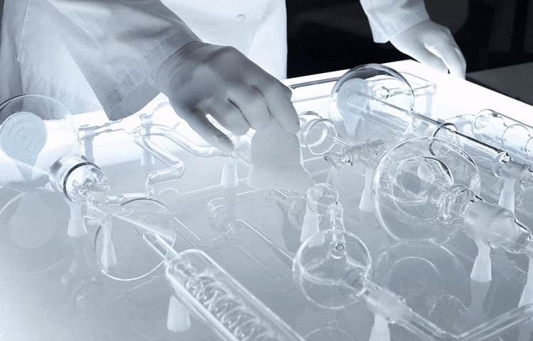 Hands wearing gloves manipulate glass laboratory equipment on a lighted surface.