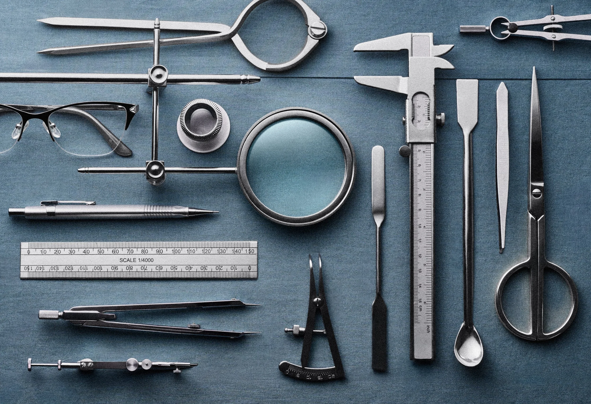 A collection of medical and scientific tools including a pair of glasses, a pair of scissors, a mirror, a caliper, a pen, a ruler, and various precision instruments laid out on a blue surface.