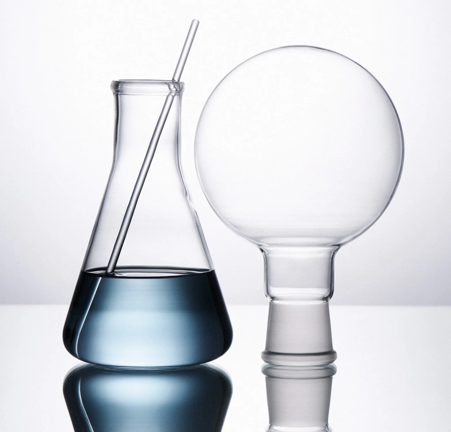 Glass lab equipment, including a conical flask with a dark blue liquid and a glass sphere with an opening at the bottom, on a reflective surface.