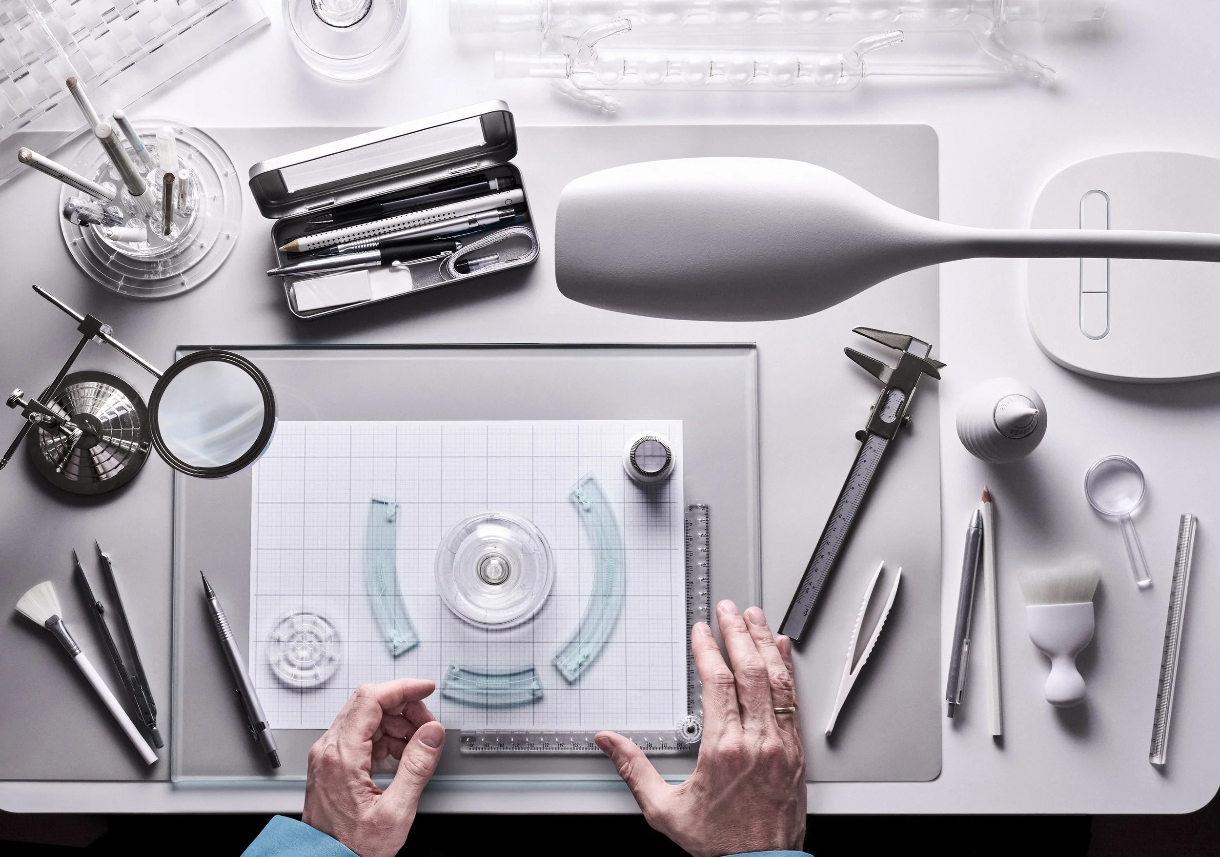 Top-down view of a scientist's workspace with glass laboratory equipment, tools, and a microscope, with hands assembling glassware on a cutting mat.