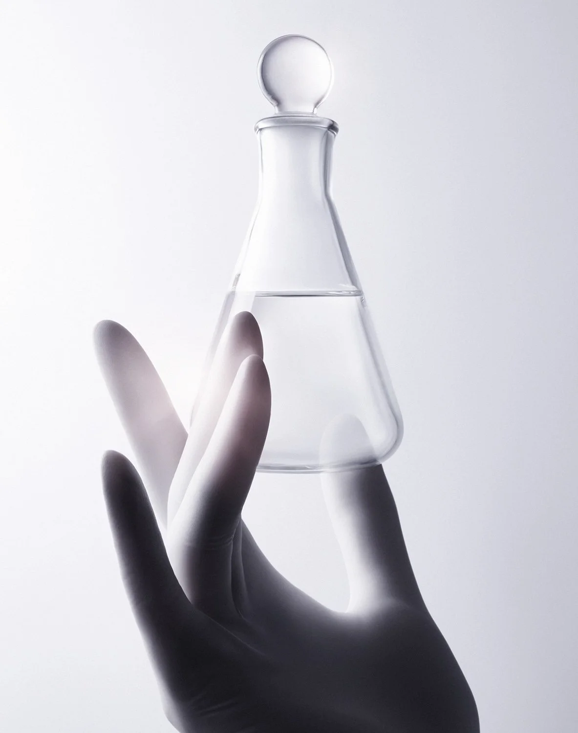 A black-and-white photo of a hand wearing a glove holding a glass Erlenmeyer flask filled with a clear liquid.