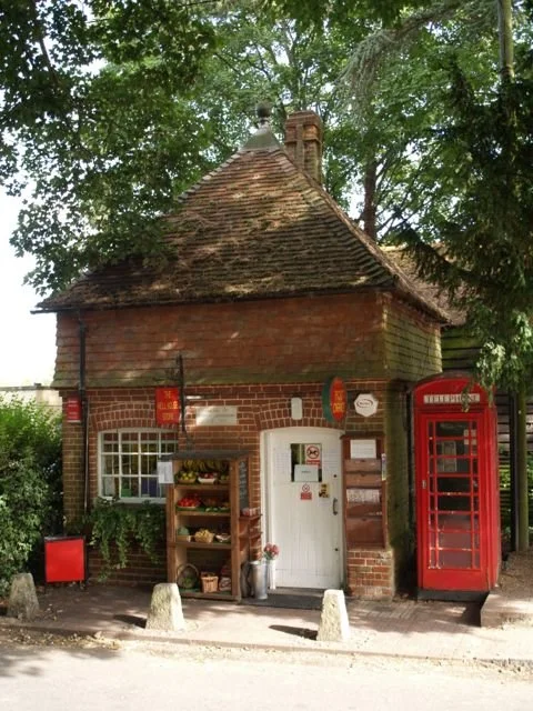 A small red brick building with a sloped roof, white door, and a red British telephone box on the right side, surrounded by trees.