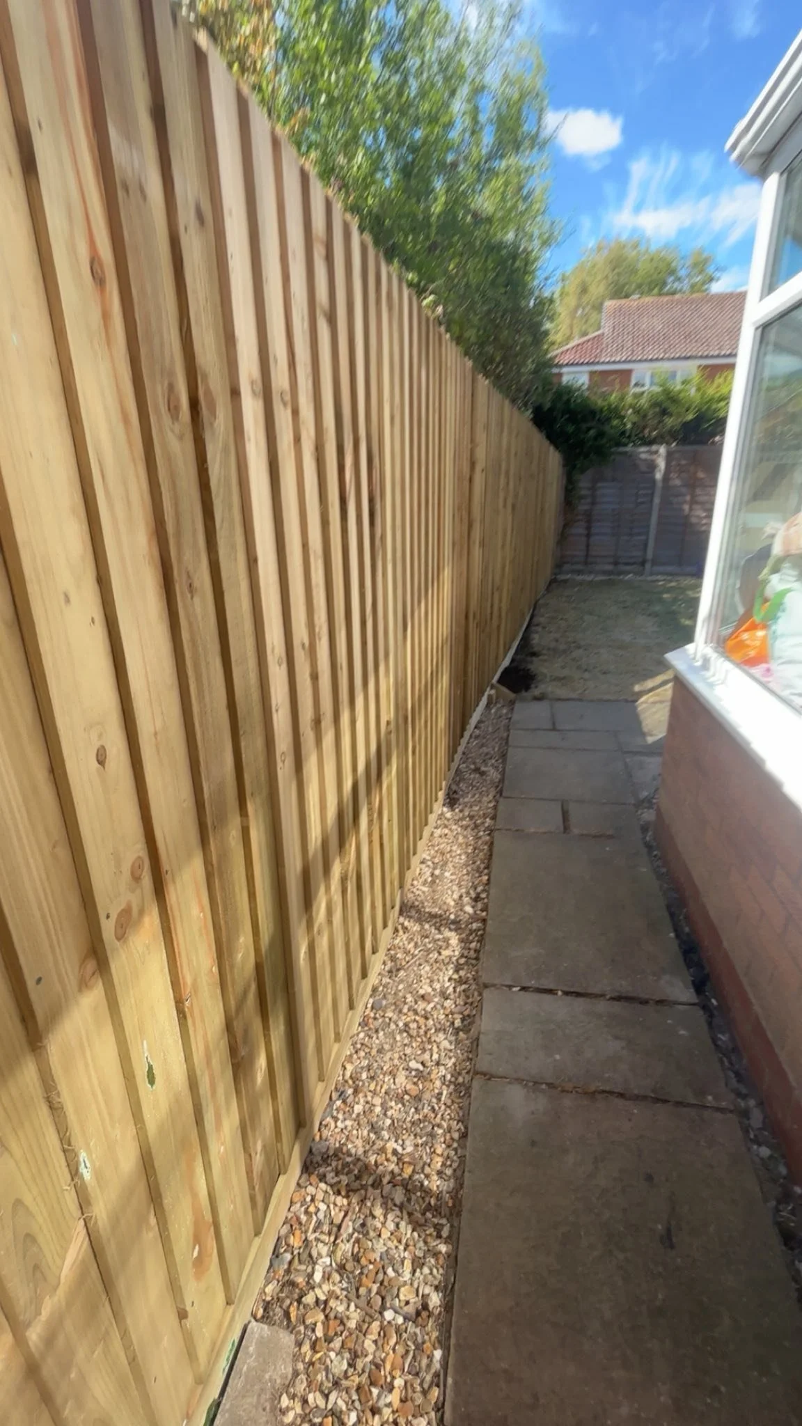 A narrow outdoor side yard with a new wooden fence, a paved path, and a garden area with gravel. A house with a window is on the right, and a blue sky with clouds is overhead.