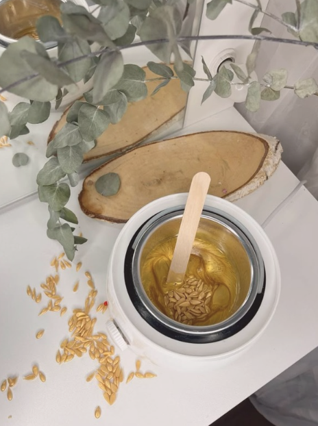 Une bouilloire électrique contenant des graines de lin dans de l'huile, avec des graines de lin dispersées sur la surface blanche et des éléments de décoration en bois et en feuilles vertes dans l'arrière-plan.