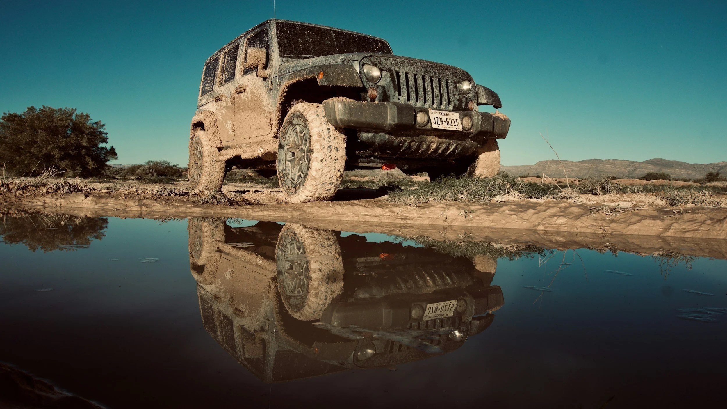 A muddy Jeep Rubicon parked next to a water puddle on a dirt trail in a desert landscape, with its reflection visible in the water.