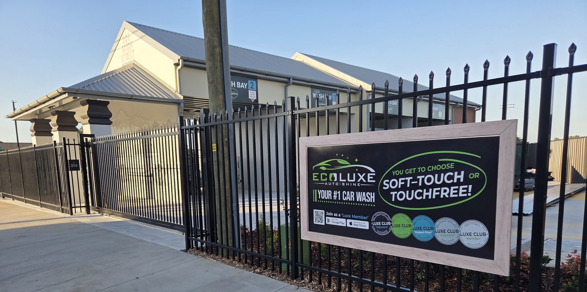 Sign outside EcoLuxe Auto Shine car wash offering touchless and soft-touch wash options, with a gated entrance and a building in the background.