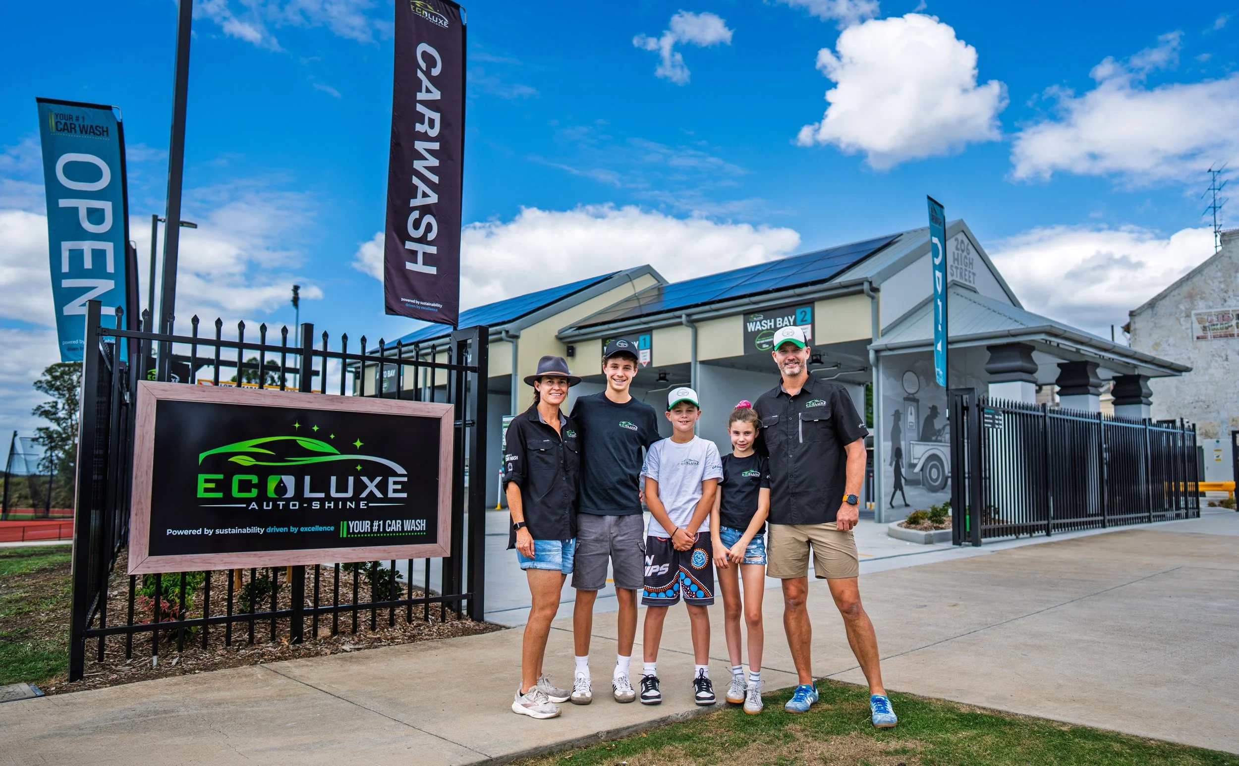 A group of five people, including two adults and three children, standing in front of a car wash facility called EcoLuxe Auto-Shine on a sunny day with blue skies and clouds.