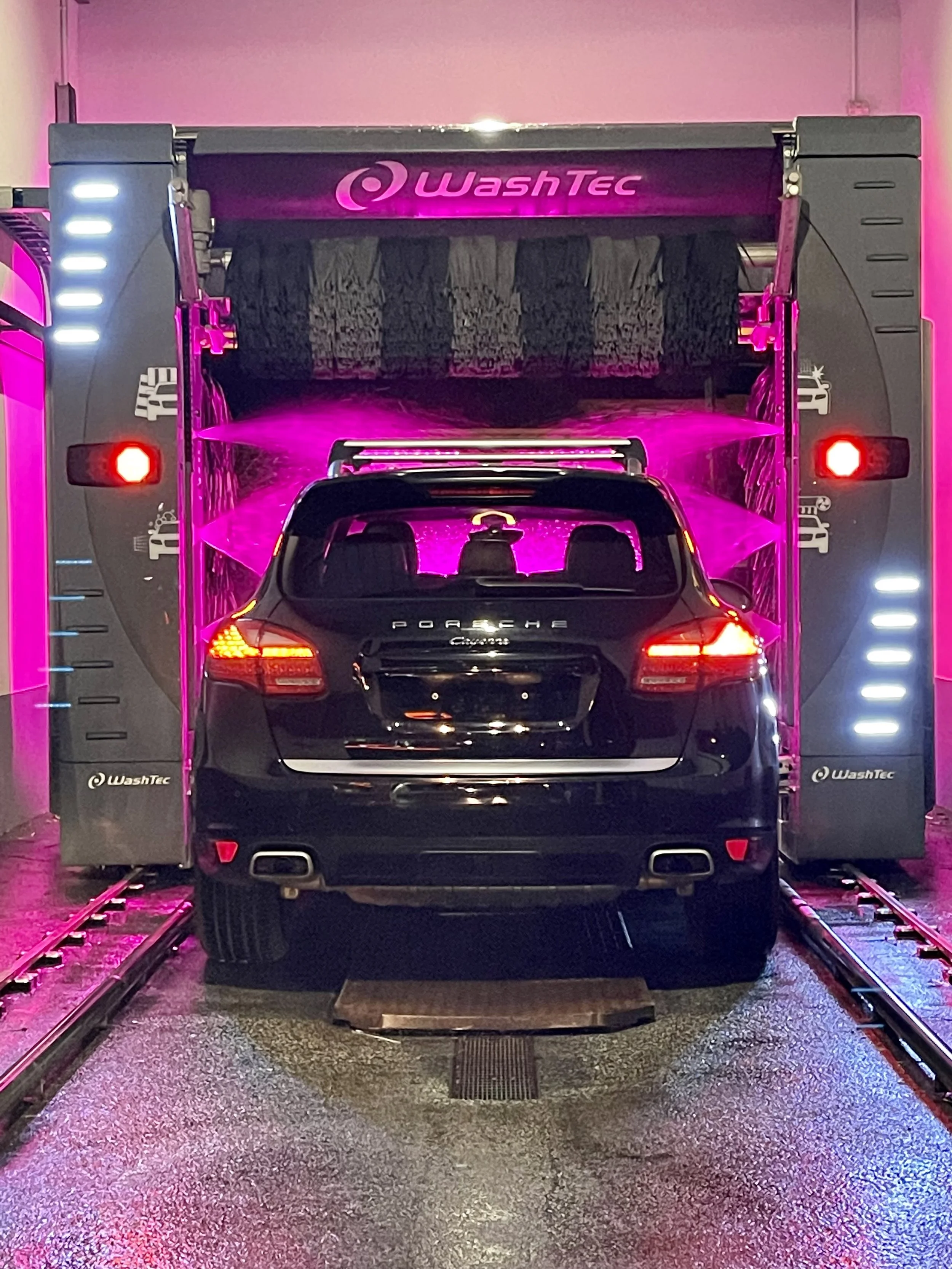 Black Porsche Cayenne inside a car wash with pink lighting and brushes.