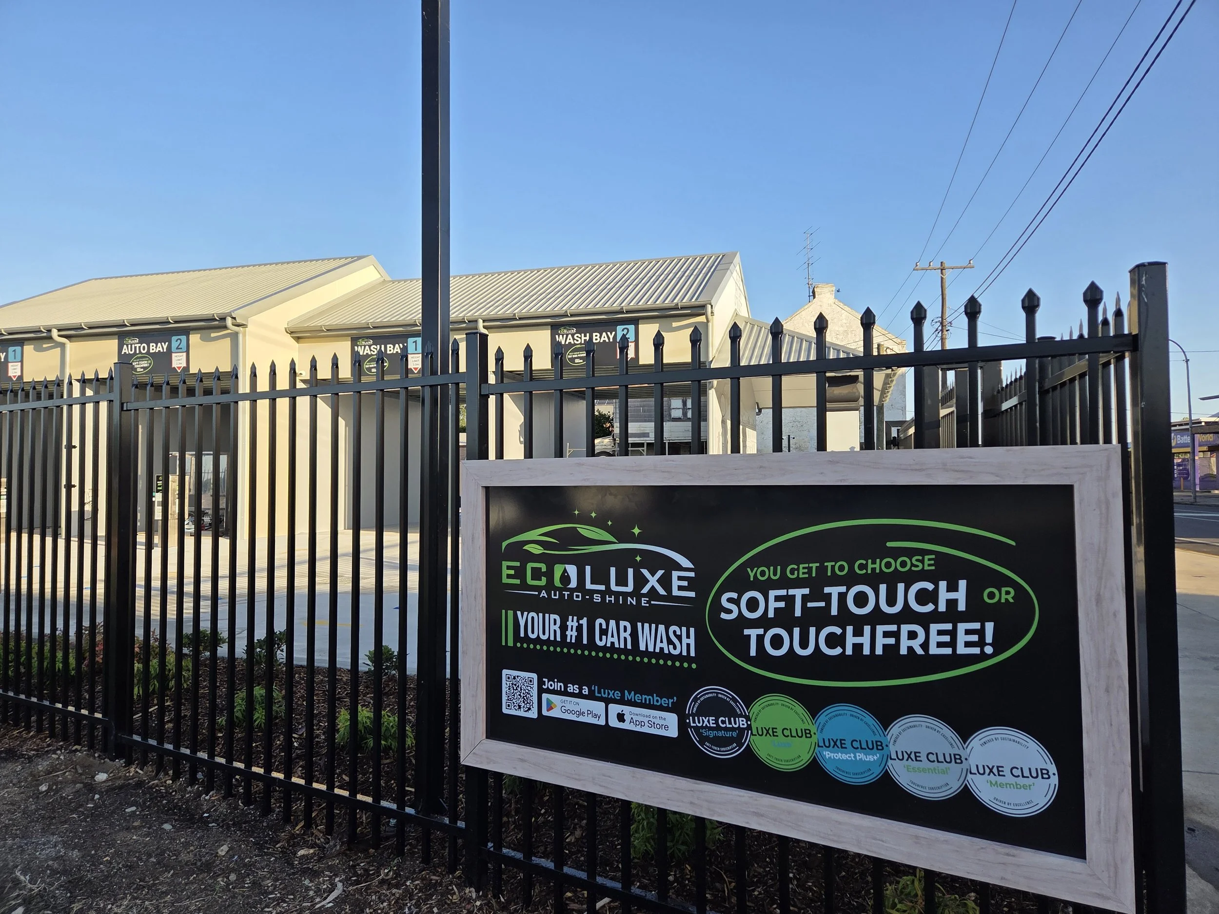 Sign outside Luxe Car Wash advertising eco-friendly automatic car wash with various membership options and a QR code, background shows a building and power lines under a clear blue sky.