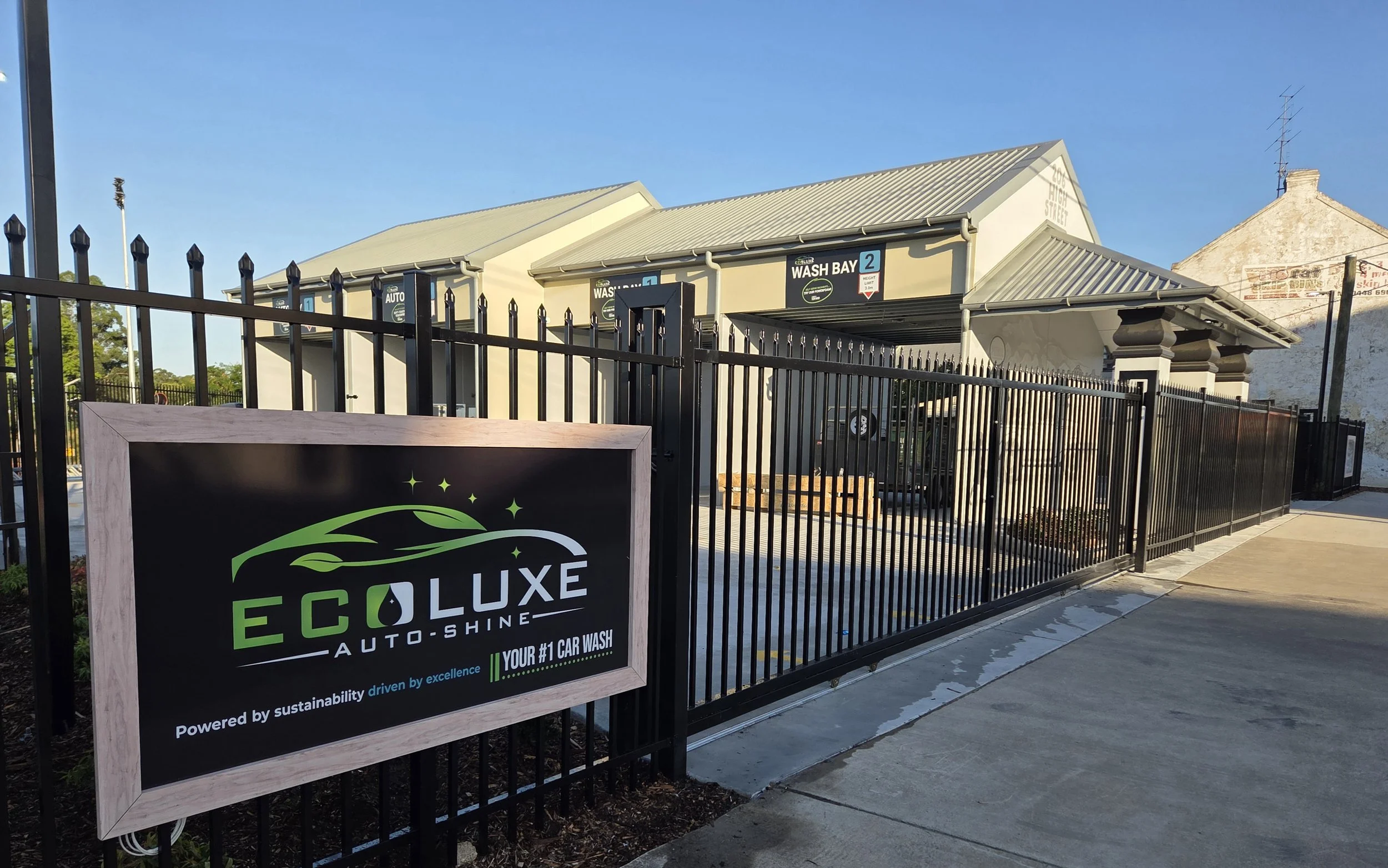 Exterior view of EcoLuxe Auto-Shine car wash with a sign in the foreground and a black metal fence surrounding the wash bays. The building has a gray, sloped roof and signs indicating wash bay two and auto services. Clear blue sky in the background.