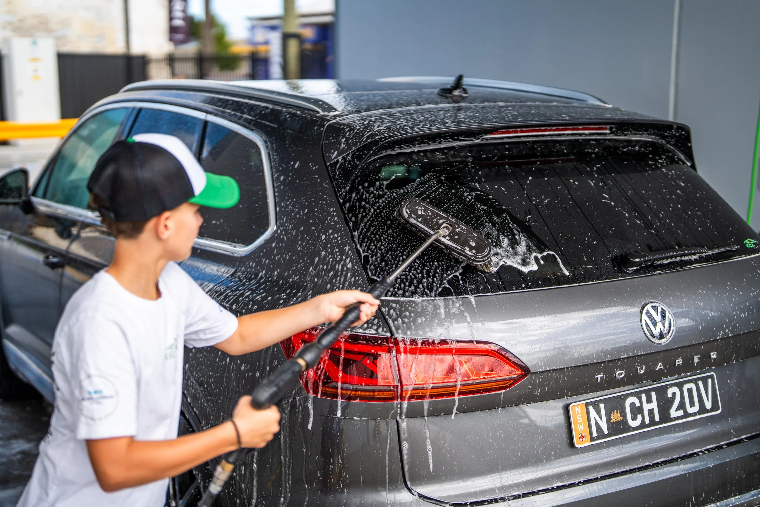 A boy washing a black Volkswagen Touareg SUV with a handheld pressure washer, soap and water on the vehicle, at an outdoor car wash station.