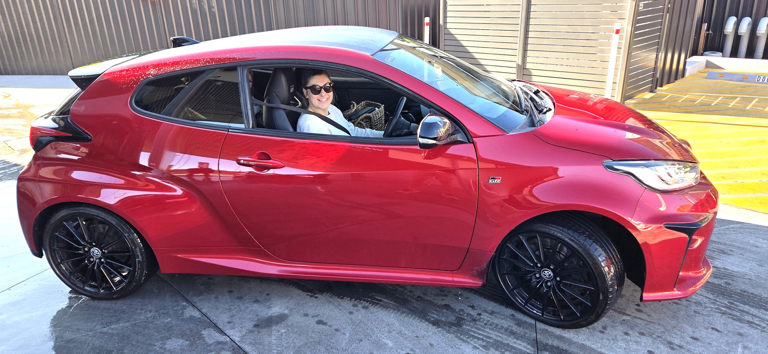 Smiling woman with dark hair and sunglasses sitting in a red sports car parked in a driveway, with a brown wooden fence and a yellow parking space in the background.