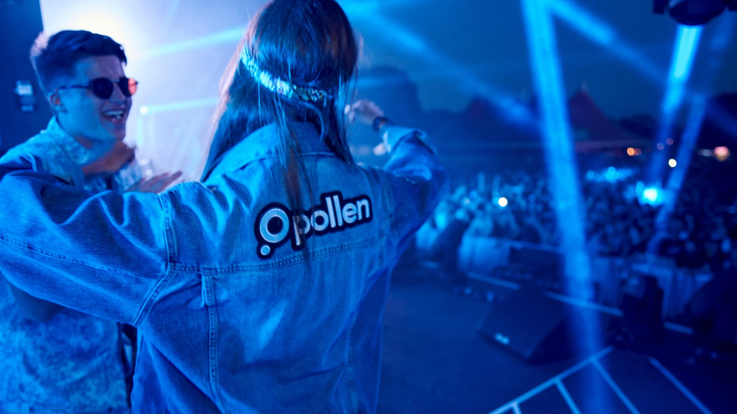 Two people at a concert or music event, one facing away wearing a denim jacket with a logo, and the other smiling and wearing sunglasses and a hoodie, with blue laser lights and a crowd in the background.