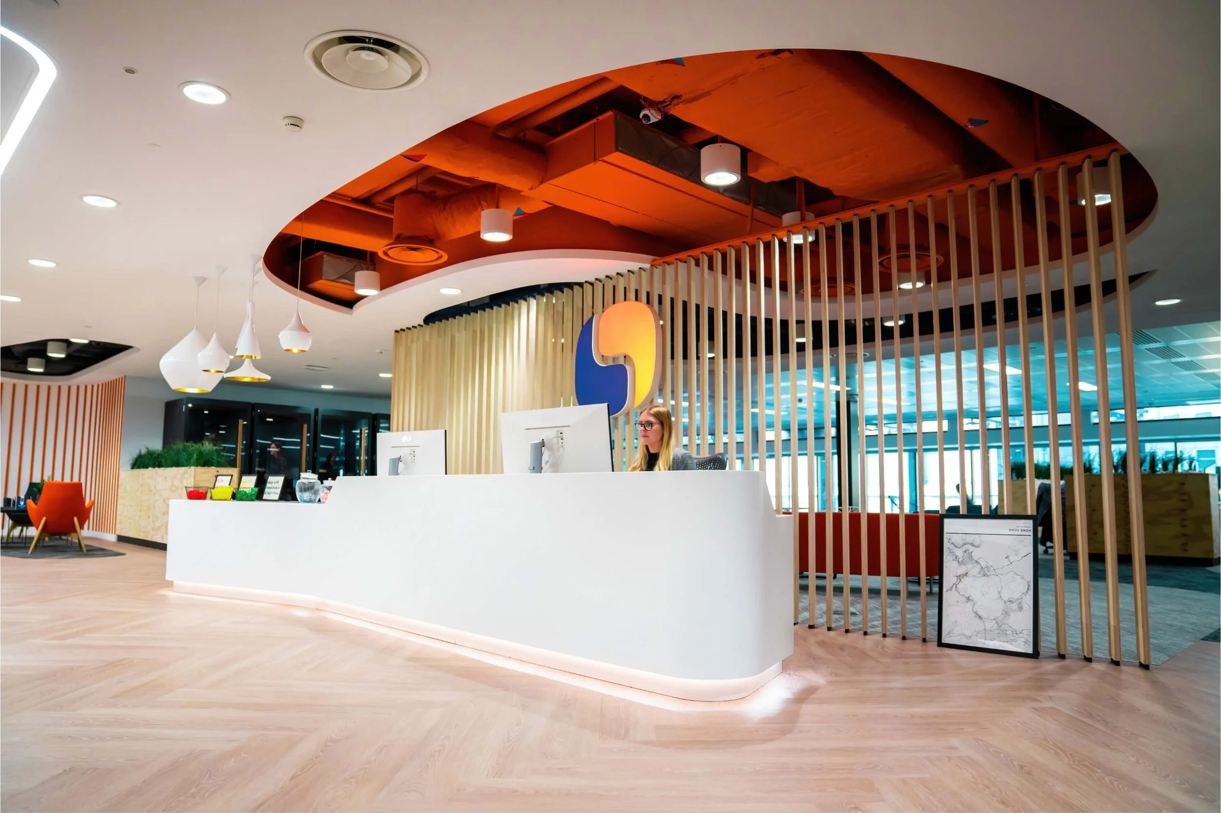 Modern office reception area with a curved white desk, a woman working on a computer, decorative wooden slats, and colorful geometric artwork on the wall.