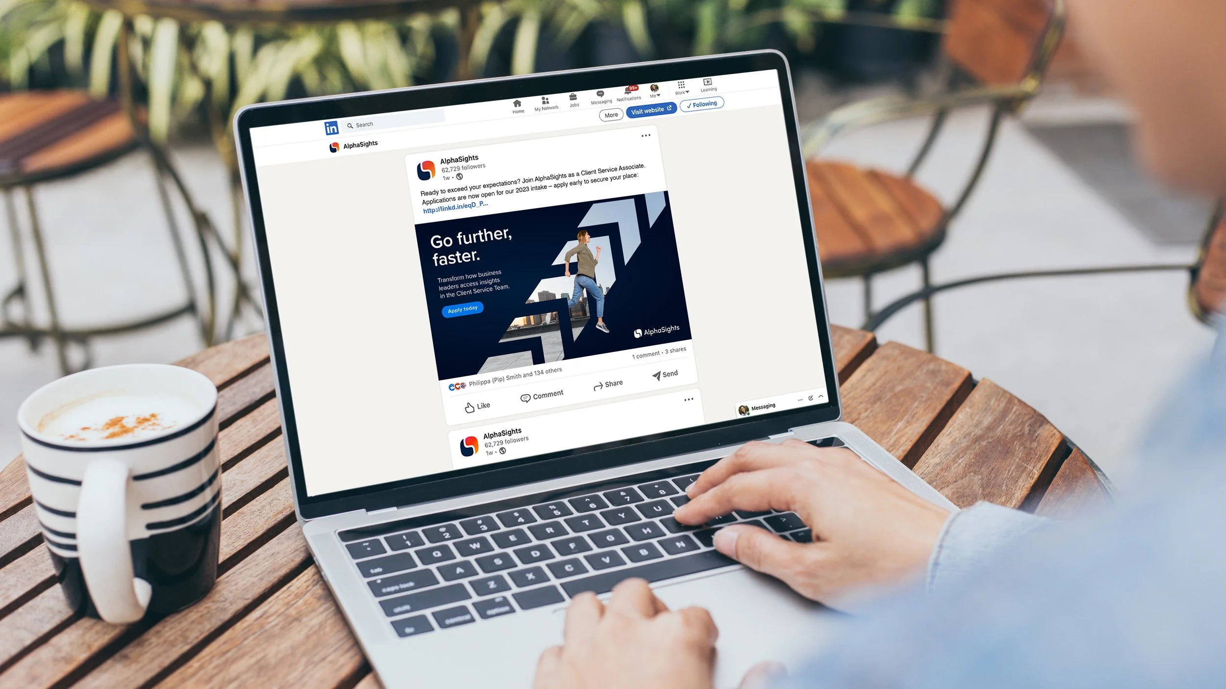 Person using a laptop at an outdoor wooden table, viewing a LinkedIn post for AlphaSights, with a black and white striped coffee mug nearby and green foliage in the background.