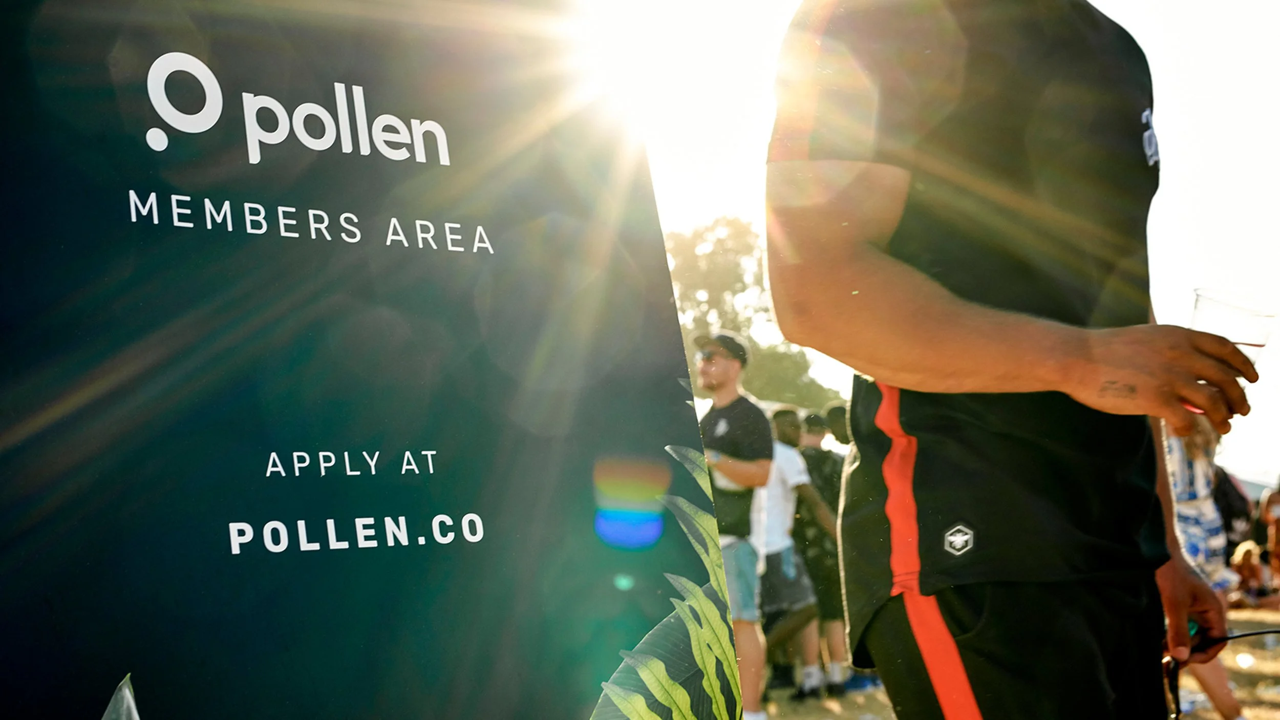 A sign that reads 'pollen MEMBERS AREA APPLY AT POLLEN.CO' with people gathered outdoors in the background, with sunlight shining and a person holding a plastic cup nearby.