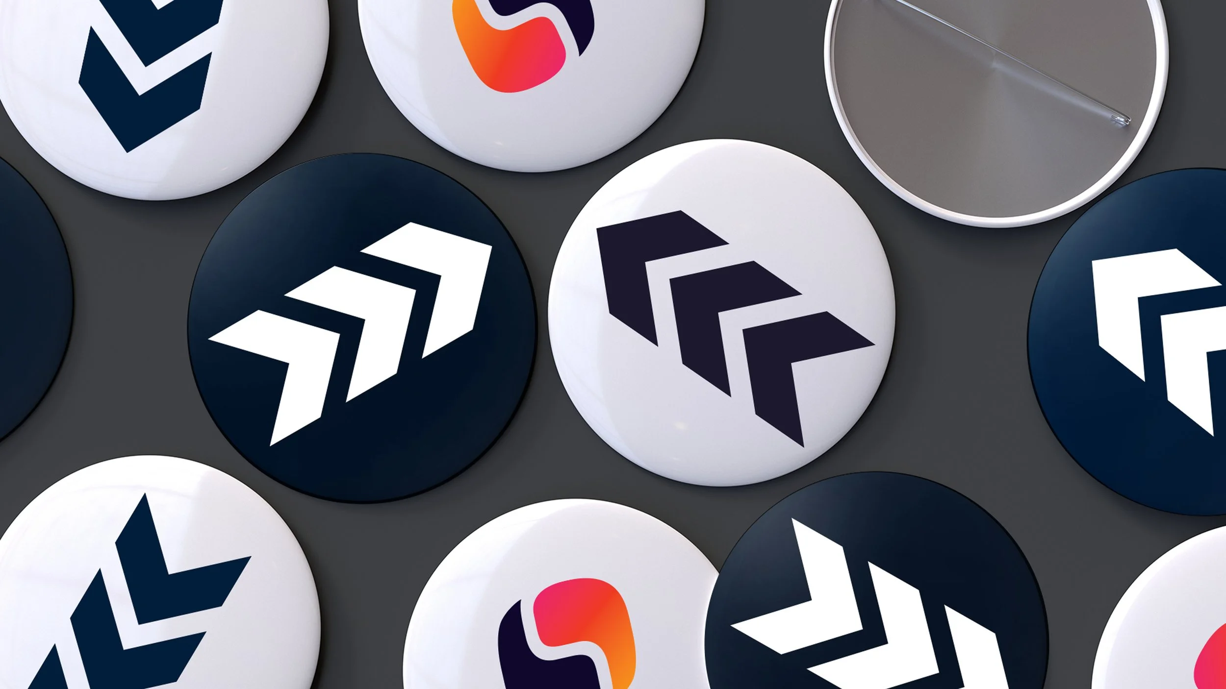 Multiple round badges with various logos, some featuring arrows or abstract shapes, placed on a dark surface, including a visible pin-back badge with a clear safety pin on the back.