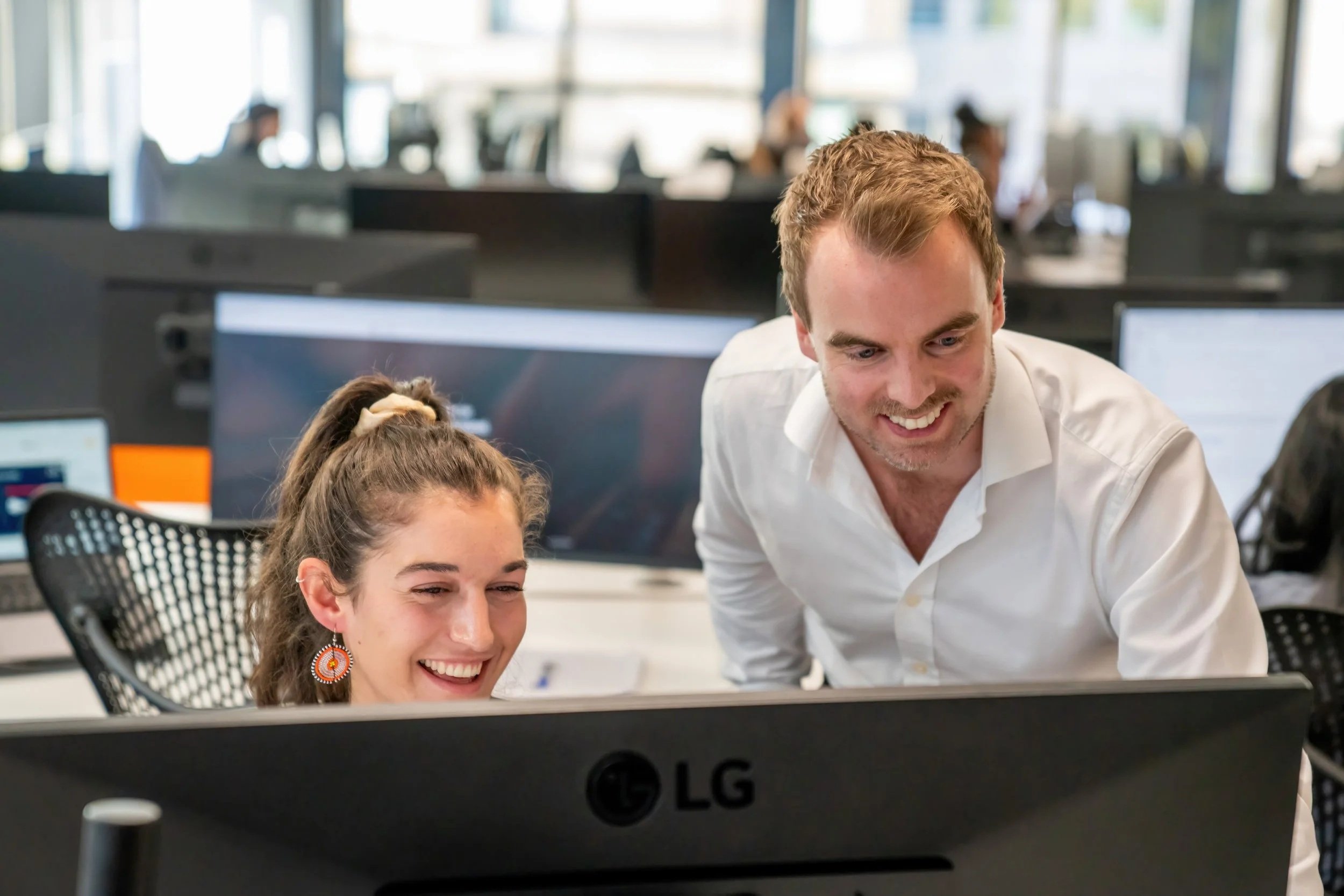 Grant art directing a colleague at a desk in an office with multiple computer monitors. They are smiling and looking at a computer screen.
