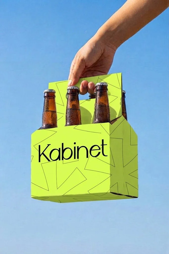 A hand holding a green six-pack of bottled beverages labeled 'Kabinet' against a clear blue sky.
