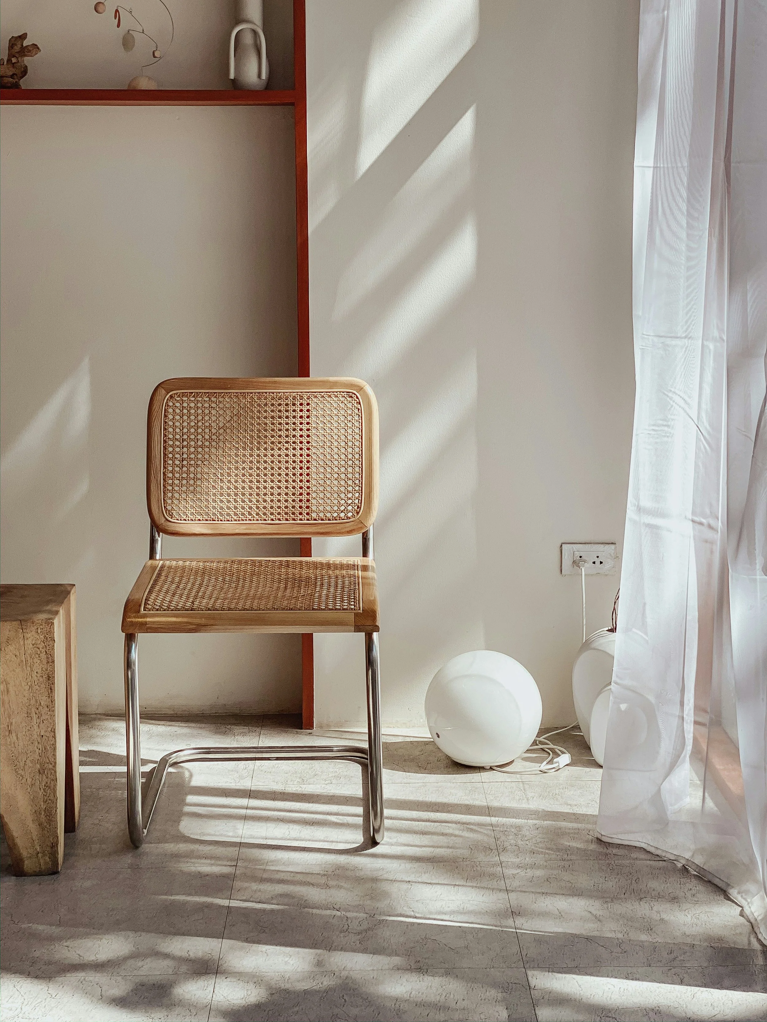 A minimalist interior corner with a wooden chair with a woven rattan seat and backrest, a small wooden side table, a white spherical lamp on the floor, and sheer white curtains letting in sunlight creating shadows on the wall and floor.
