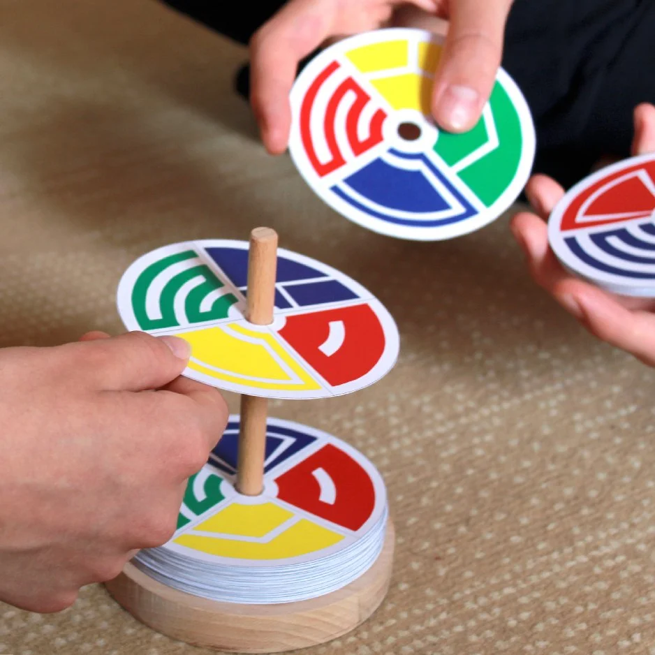 People playing a colorful board game with circular pieces that have segments in red, yellow, green, and blue. The game pieces are being stacked on wooden rods in a wooden game stand. Radovi polaznika programa priprema za Studij dizajna.