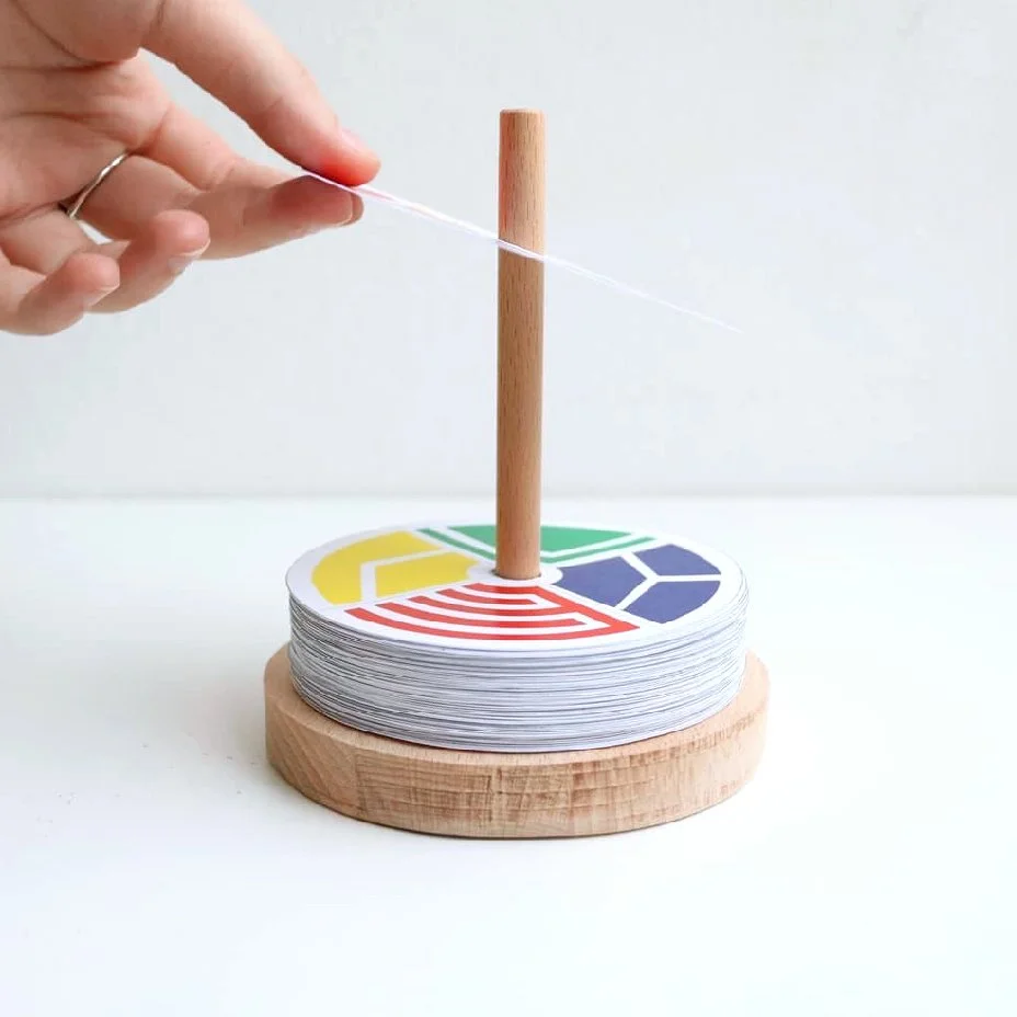 A person feeds a string through a wooden stick on top of a large stack of color-coded cards, which lay on a round wooden base.