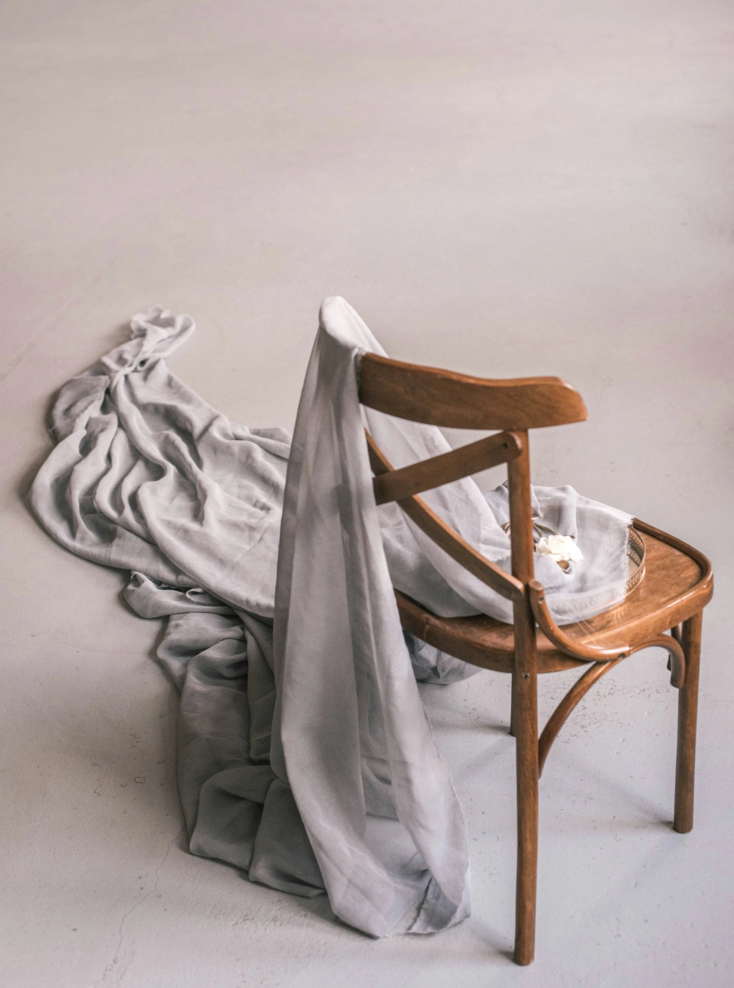 An empty wooden chair with a white sheet draped over it, a crumpled cloth on the floor nearby, against a plain beige wall.