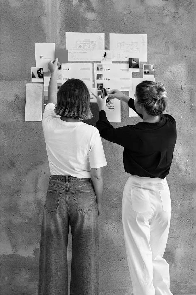 Two women standing in front of a wall with various papers, photographs, and sketches attached, discussing or working on a project.