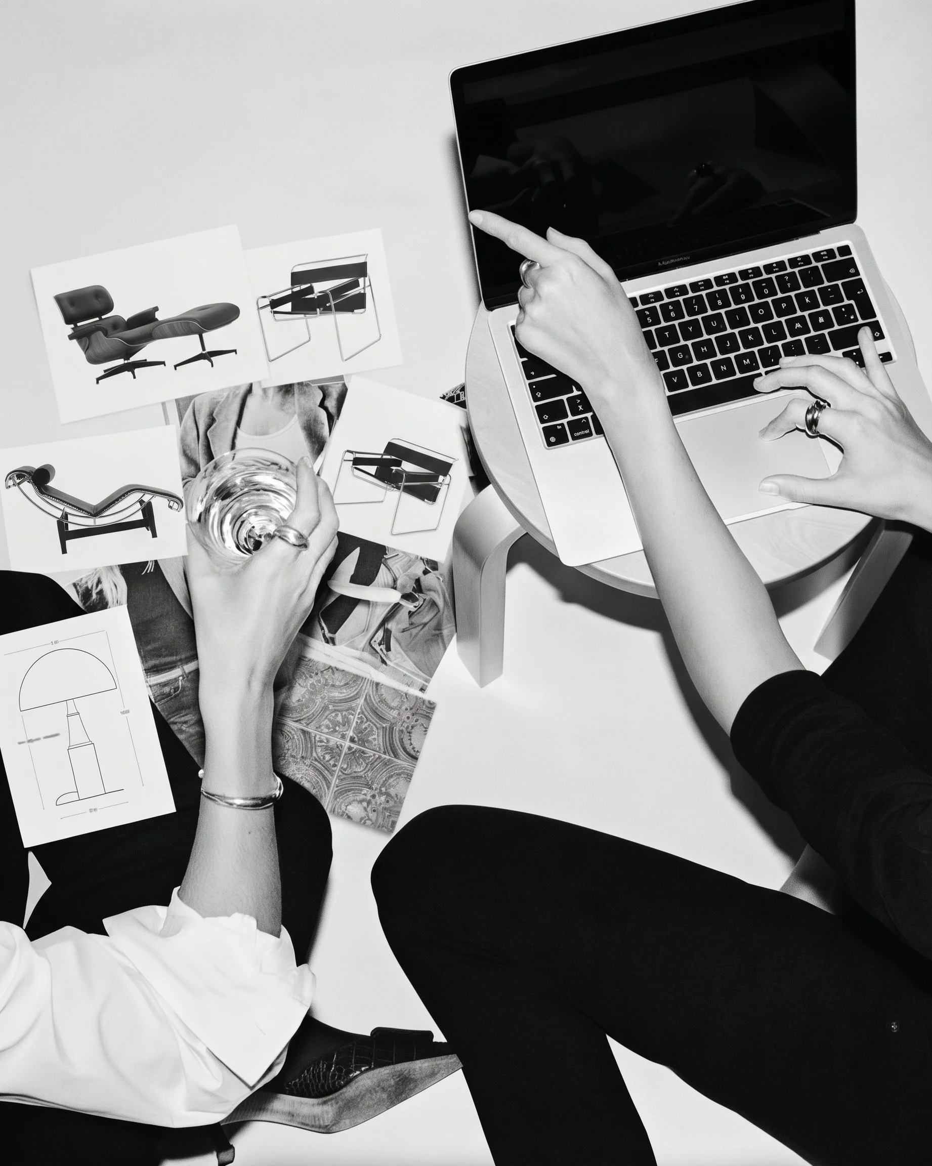 Black and white photo of two people sitting at a table, reviewing design sketches and fabric swatches. One person is pointing at a laptop, while the other is holding a glass of water.