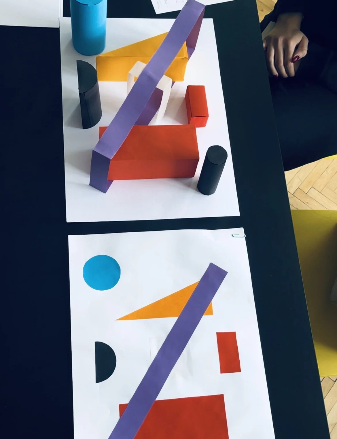 Radovi polaznika programa priprema za ŠPUD, Školu primjenjene umjetnosti i dizajna u Zagrebu.
Colorful geometric shapes including red, orange, purple, black, and blue objects on white paper and a table, with a person's hand visible in the background.