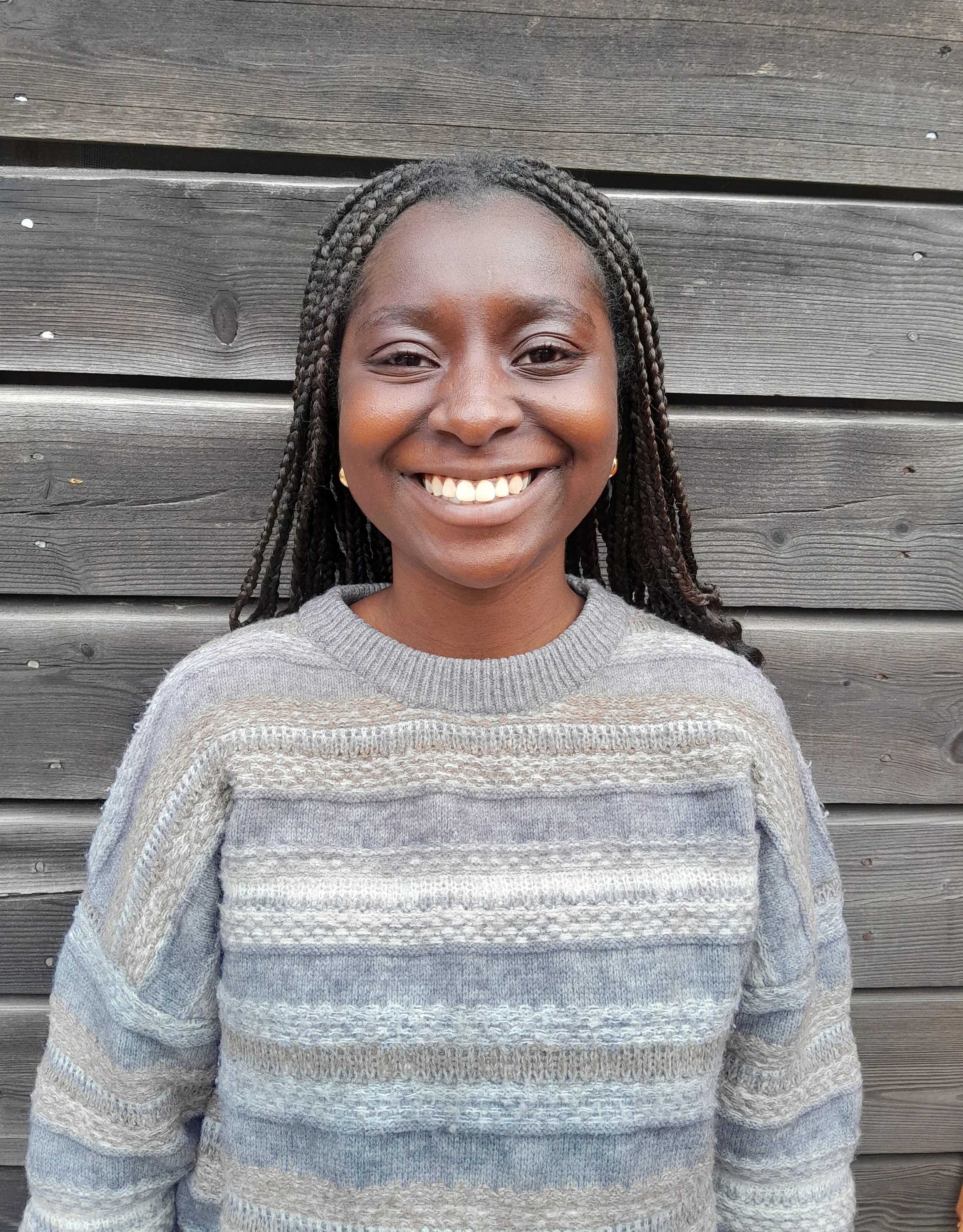 A smiling woman with braided hair in front of a wooden fence, wearing a gray patterned sweater.