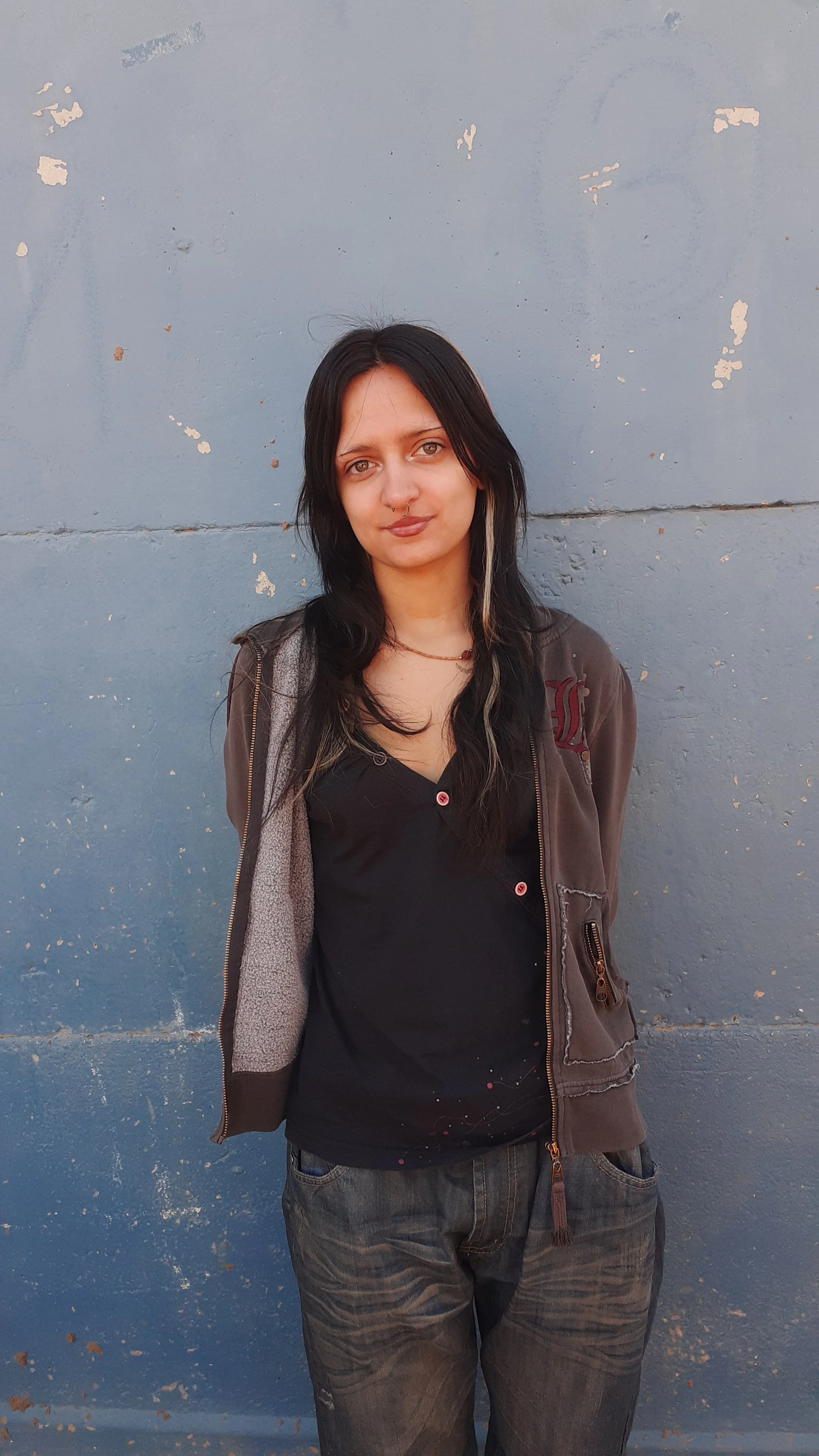 A young woman with straight black hair and a streak of blonde, wearing a dark t-shirt and a brown zip-up jacket, standing against a weathered blue wall.