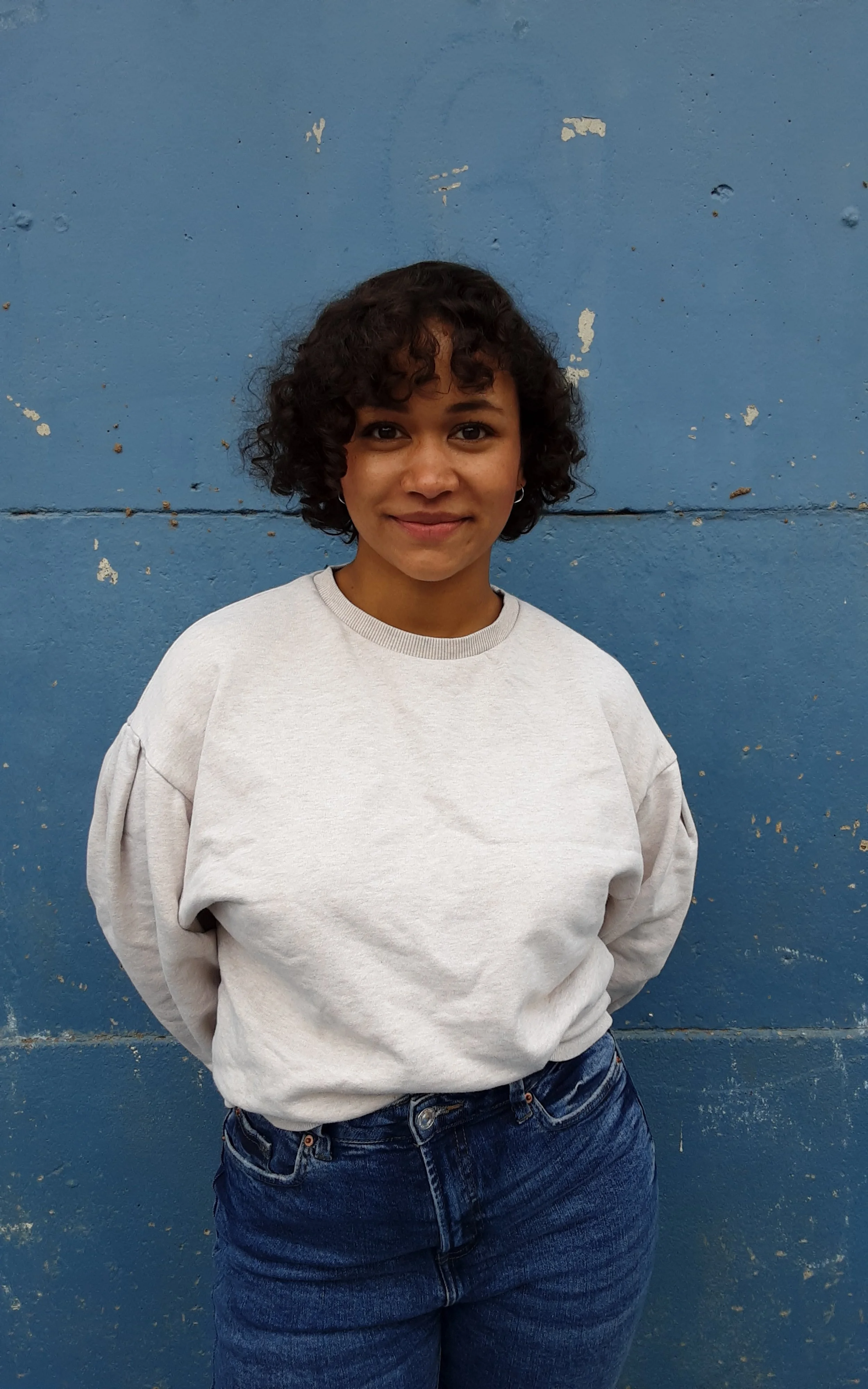 A young woman with curly dark hair smiling, standing in front of a blue wall.