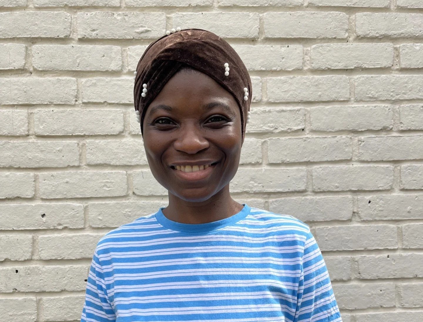 A smiling woman with dark skin, wearing a brown headwrap decorated with white pearls, a blue and white striped t-shirt, standing against a beige brick wall.
