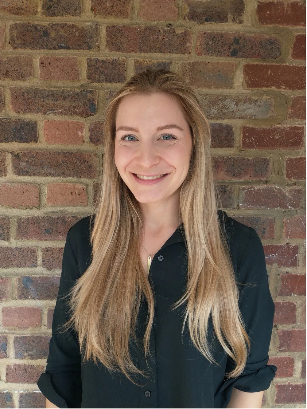 A young woman with long blonde hair, smiling, wearing a black shirt, standing in front of a brick wall.