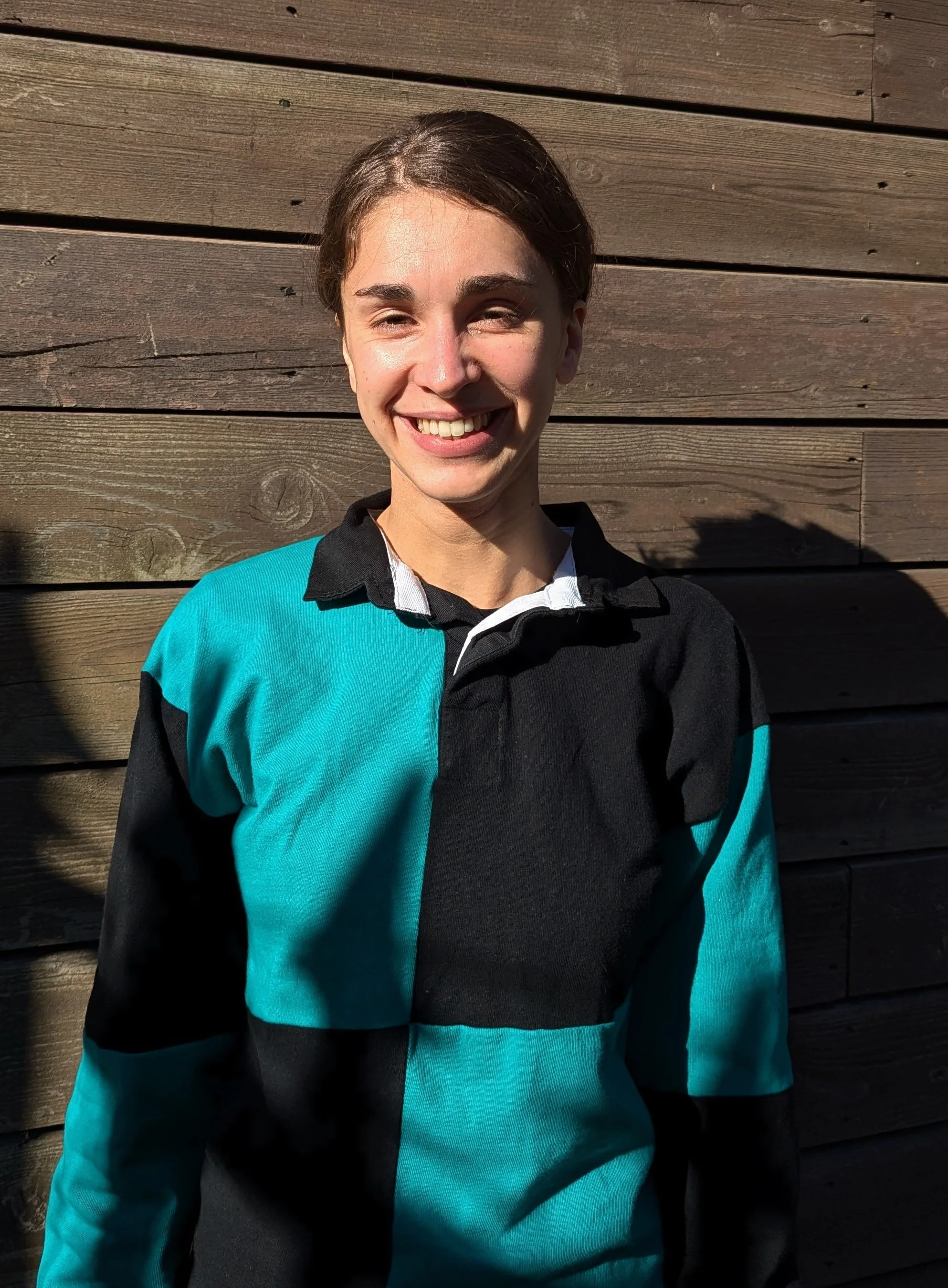 A smiling young woman with short brown hair posing outdoors in sunlight against a wooden wall, wearing a color-blocked sports jacket with black, turquoise, and white sections.