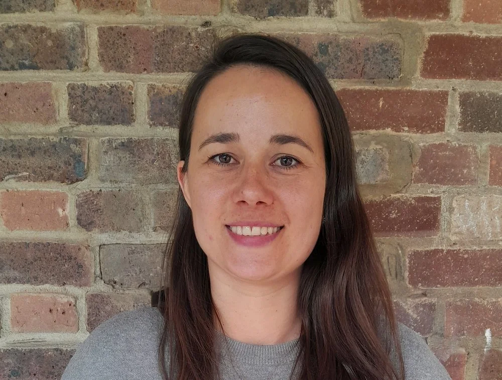 A woman with long brown hair, smiling, wearing a gray shirt, standing in front of a brick wall.