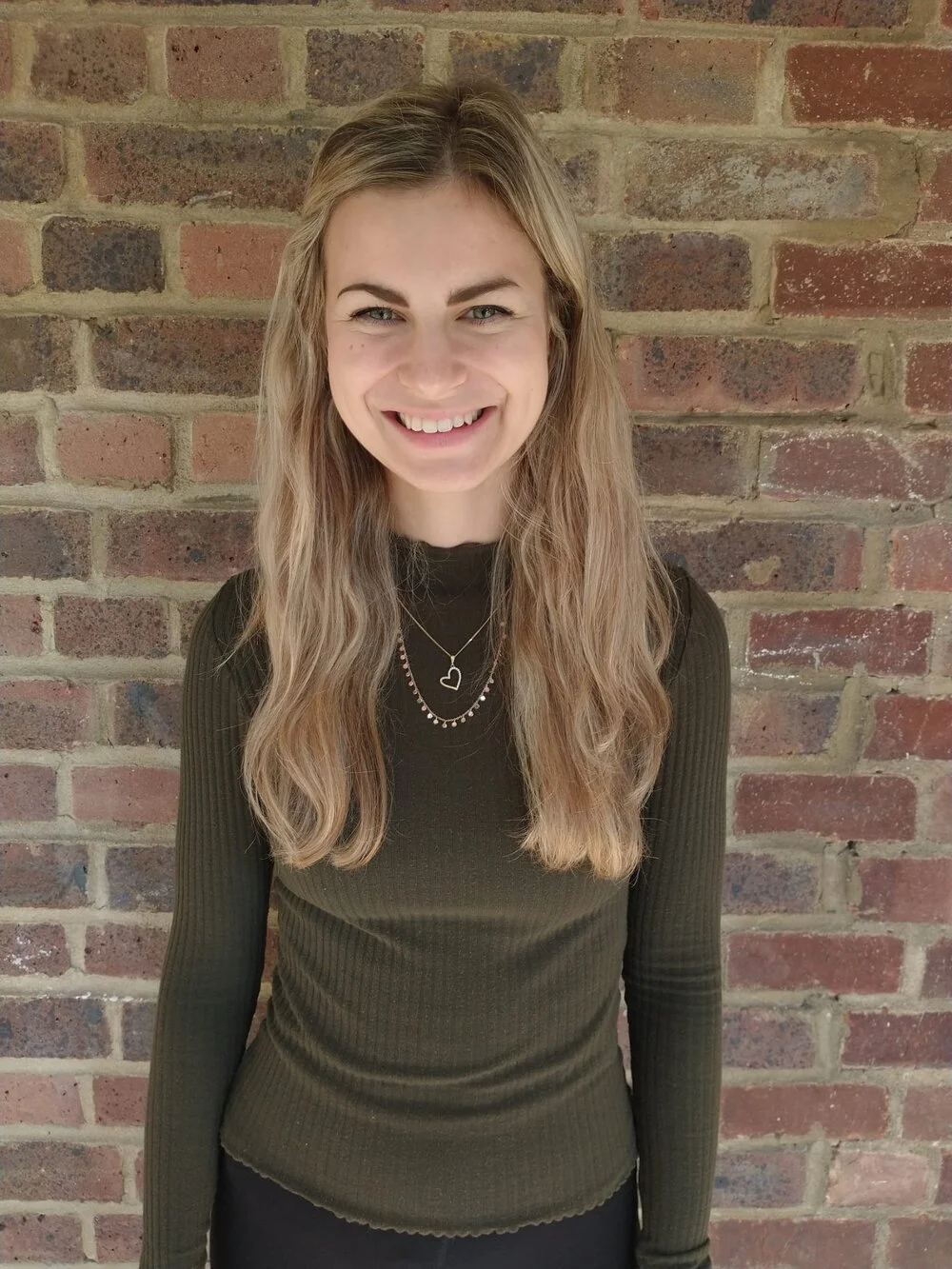 A young woman with long blonde hair smiling, standing in front of a brick wall, wearing a dark green ribbed long-sleeve top and layered necklaces.
